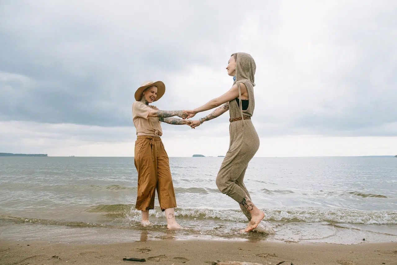 Two people holding hands and jumping in shallow water at a beach under a cloudy sky
