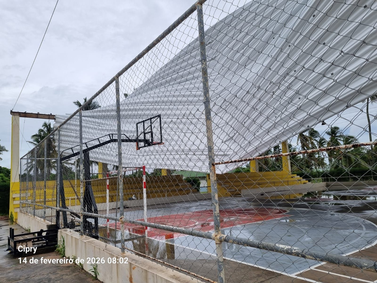 Flooded outdoor basketball court behind a chain-link fence, with a hoop and wet concrete under overcast skies.