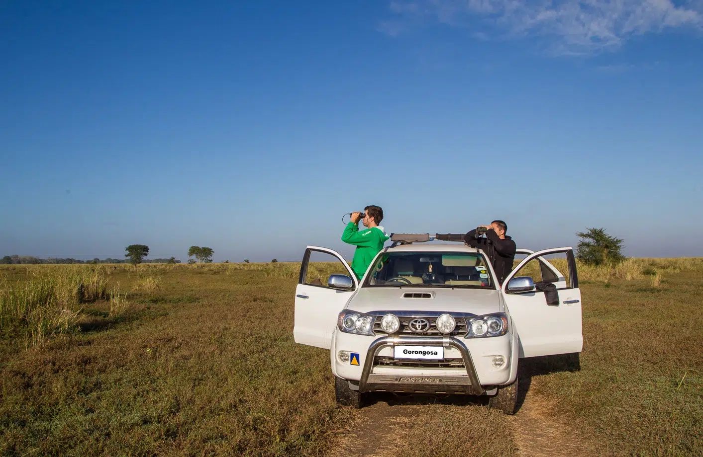 Two people standing by a white SUV in a grassy field under a clear blue sky