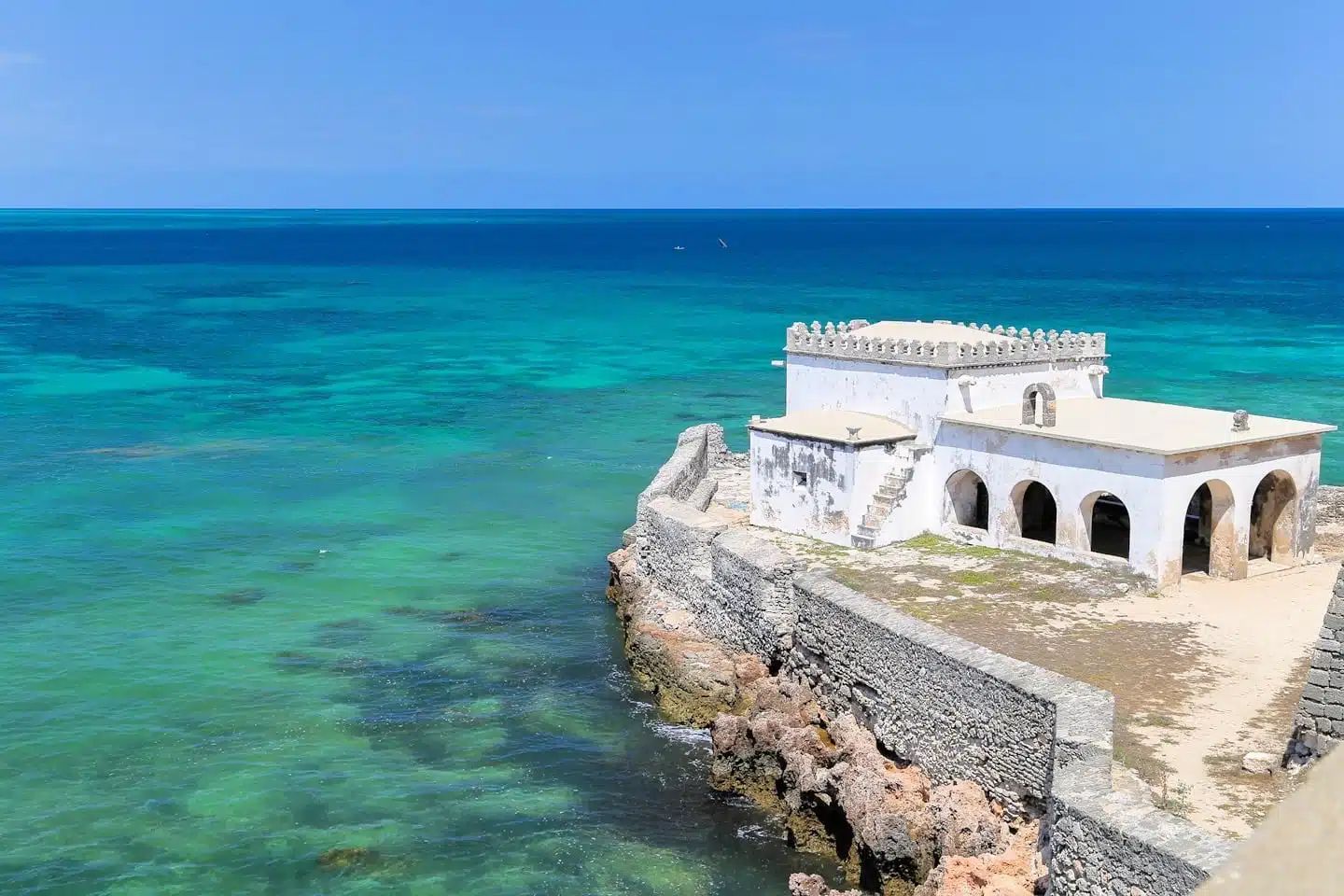 White stone building on a rocky coast above turquoise sea under a clear blue sky