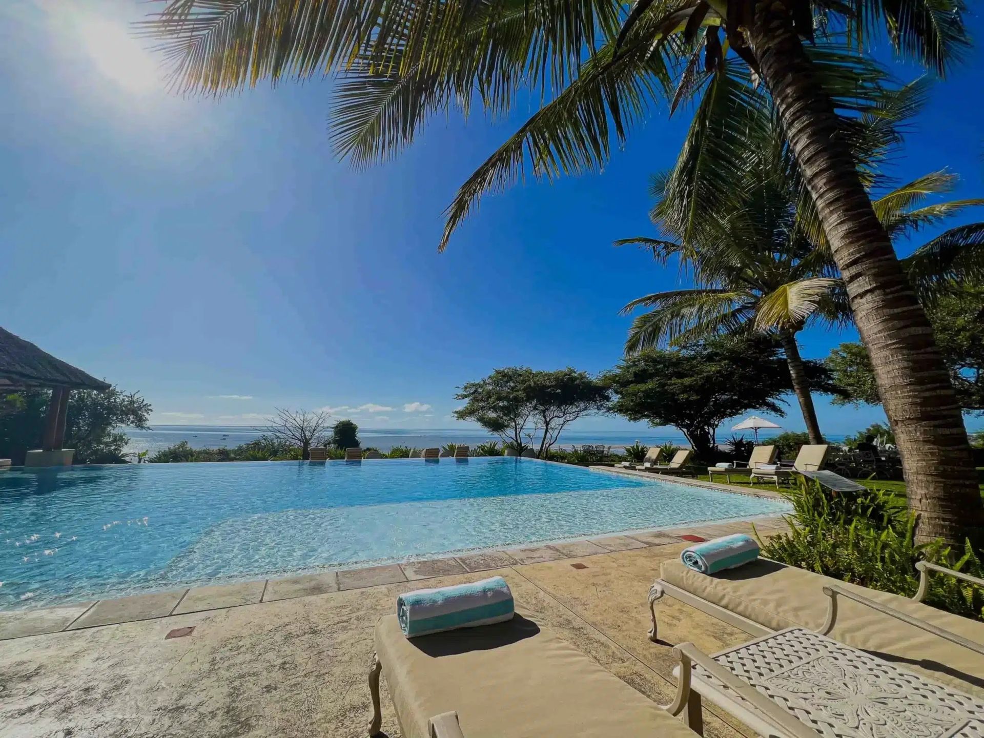 Tropical resort pool with lounge chairs, palm trees, and ocean view under a blue sky