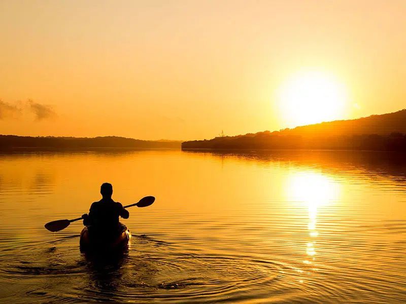 A person kayaks on calm water during a bright golden sunset, silhouette against the light.