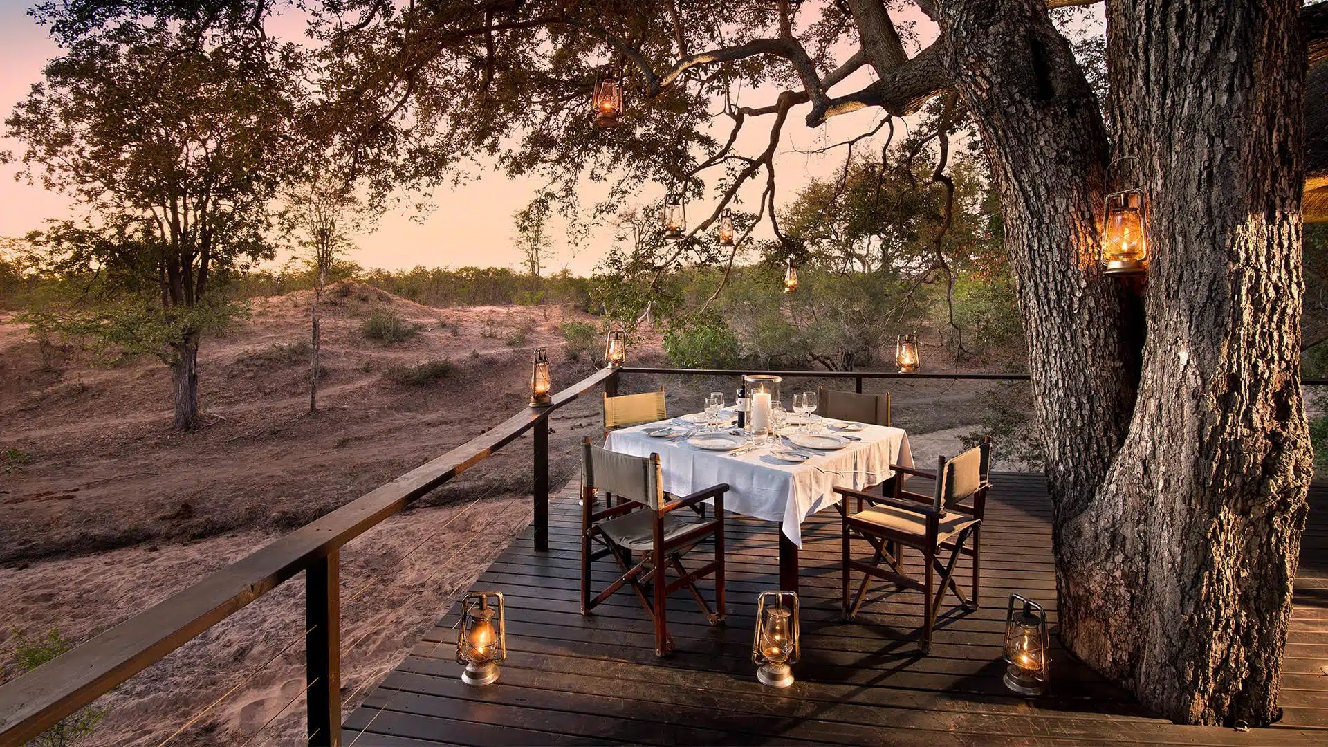 Lantern-lit outdoor dining table under trees at sunset on a wooden deck overlooking a dry landscape