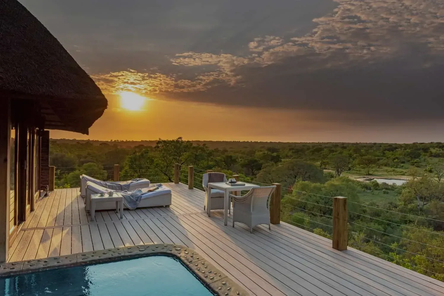 Sunset view from a deck with lounge chairs, table, and pool overlooking lush greenery