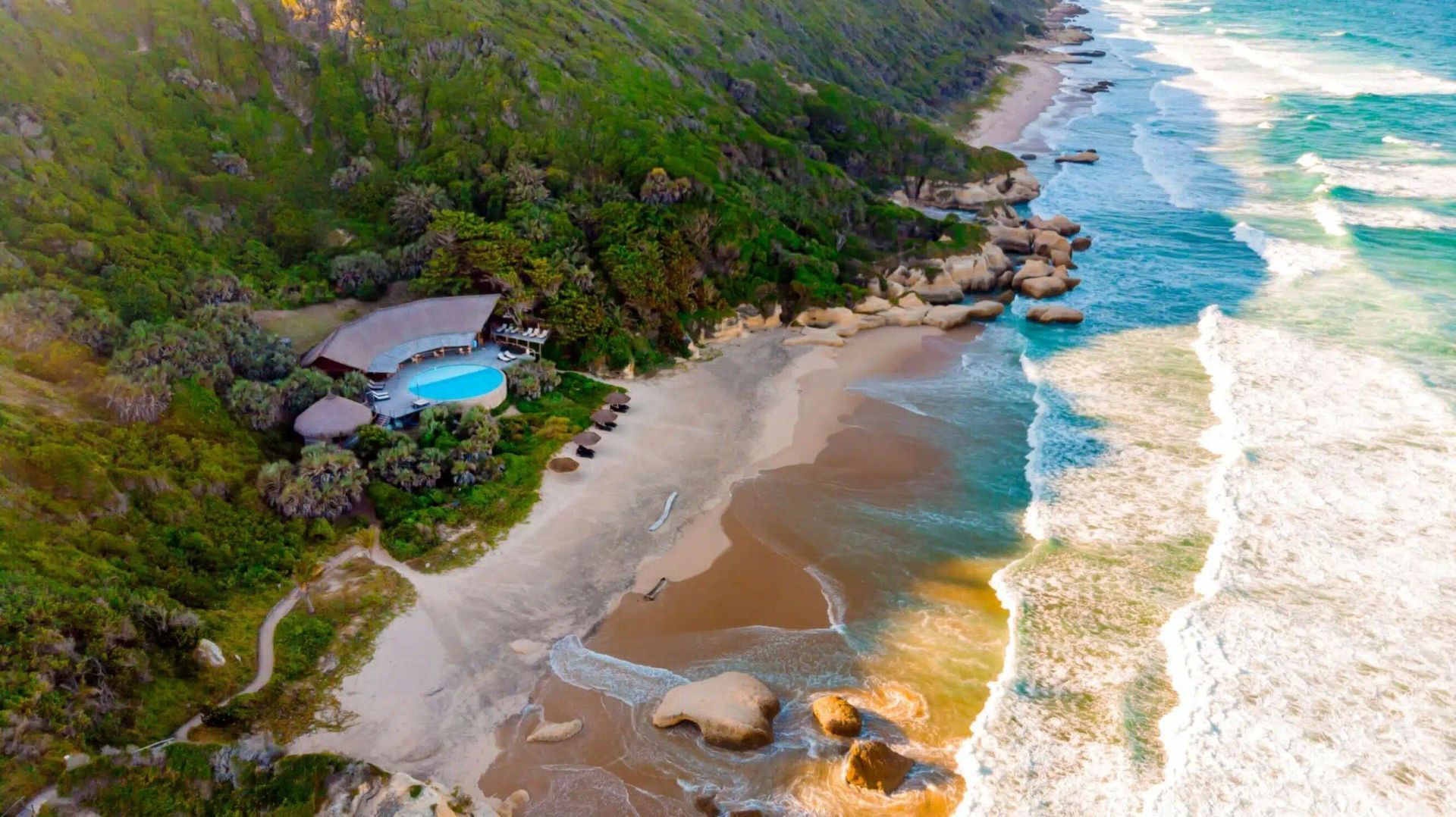 Aerial view of a secluded tropical beach with turquoise waves, golden sand, and a small hut by lush green cliffs
