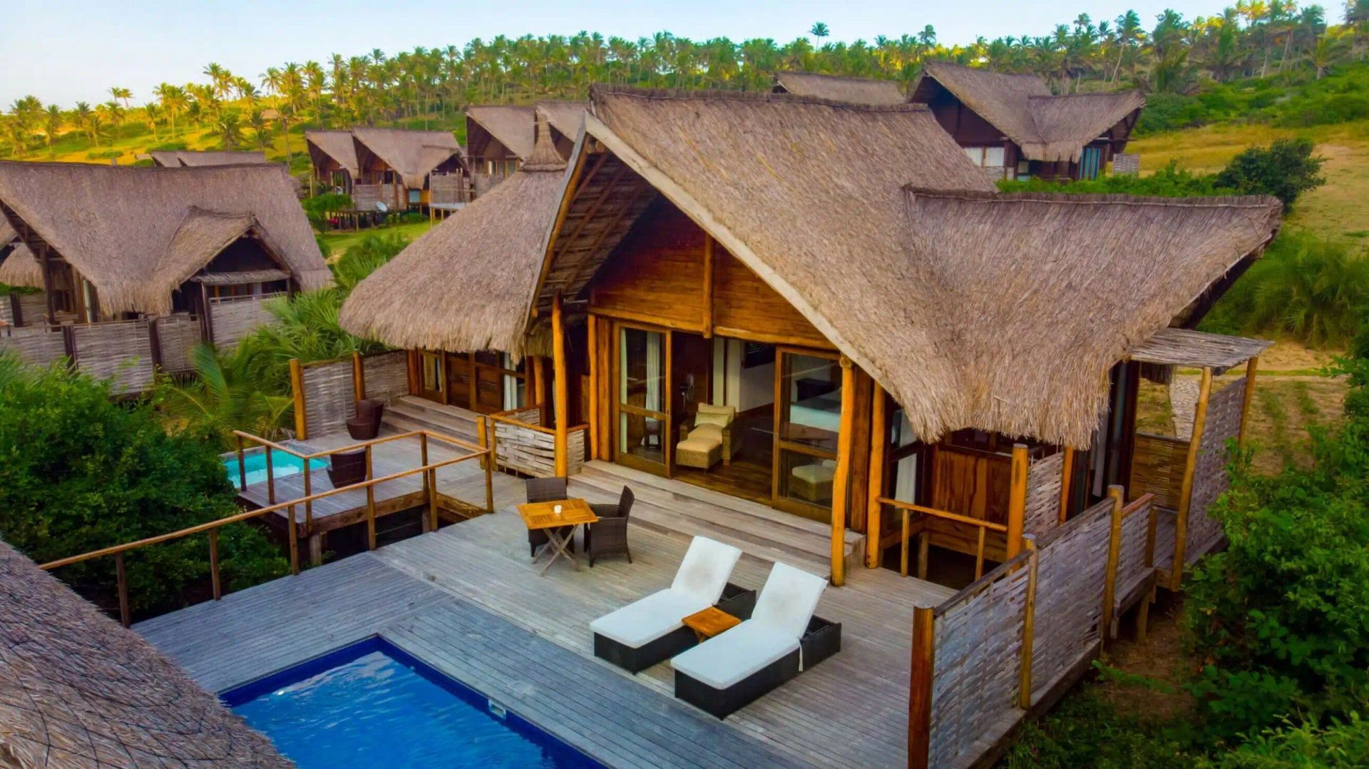 Thatched tropical villa with deck, pool, and lounge chairs in a lush green resort setting