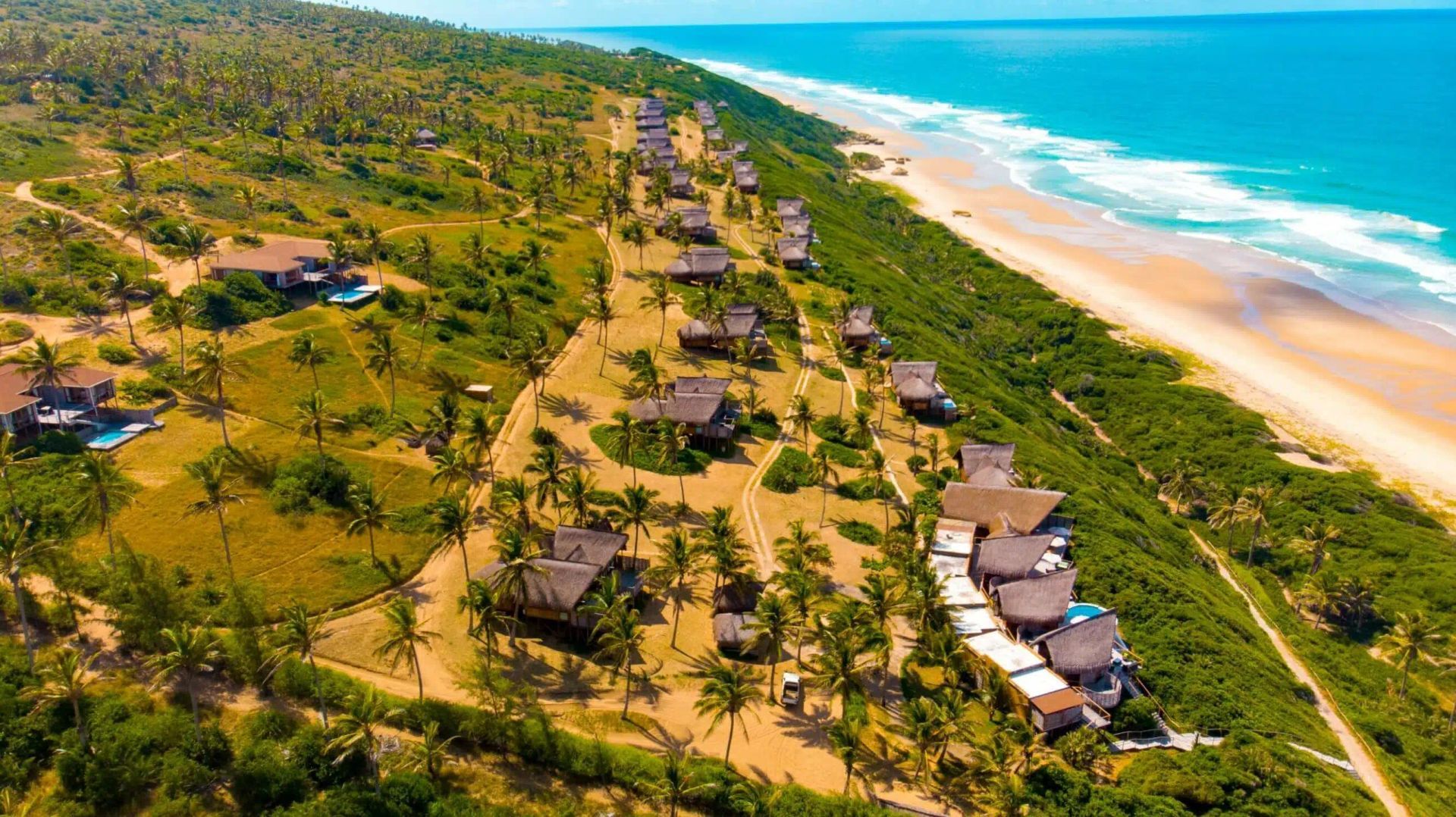 Aerial view of beachfront bungalows beside a sandy shore and turquoise ocean, bordered by dense green forest