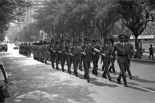 A column of soldiers in uniform marches in formation along a tree-lined city street.