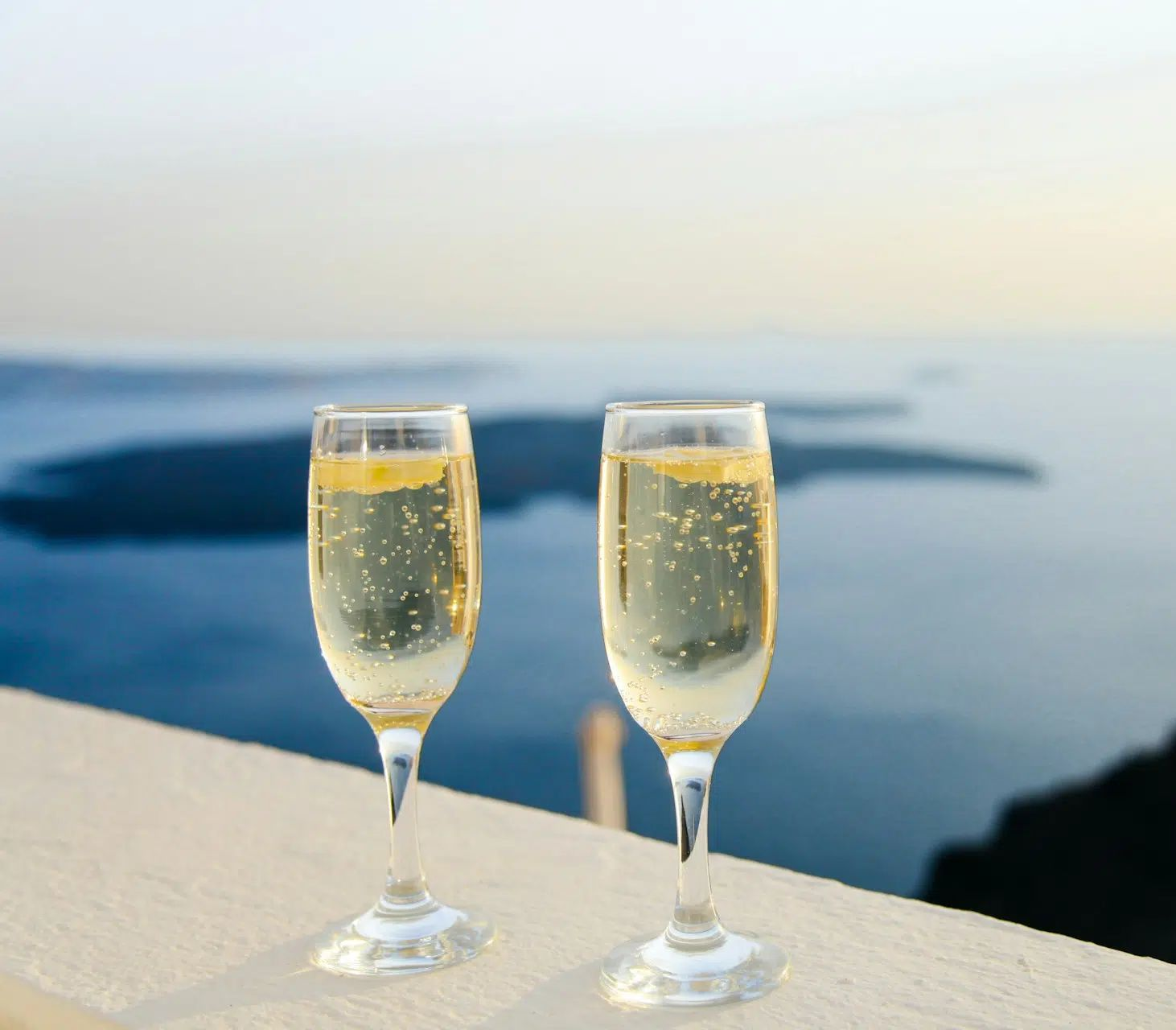 Two champagne flutes on a ledge overlooking a calm sea at sunset