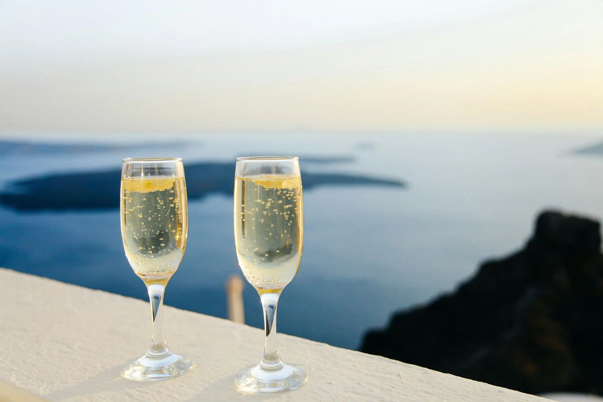 Two champagne flutes on a ledge overlooking a calm blue sea at sunset