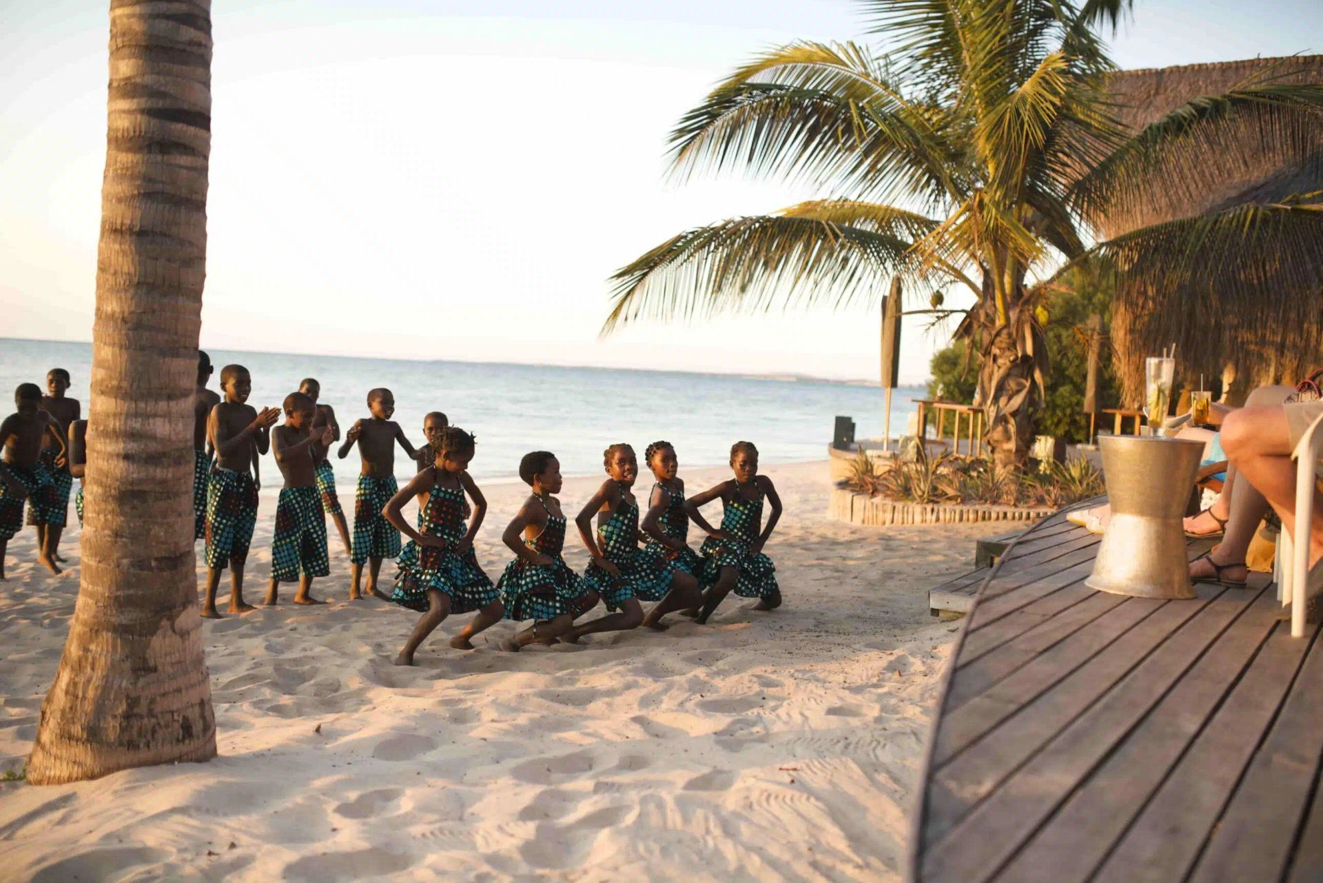 People dancing on a sandy beach at sunset beside palm trees and a wooden deck.