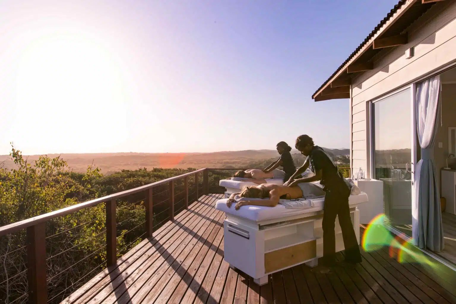 Two people receive outdoor massages on a wooden deck overlooking a sunlit landscape.