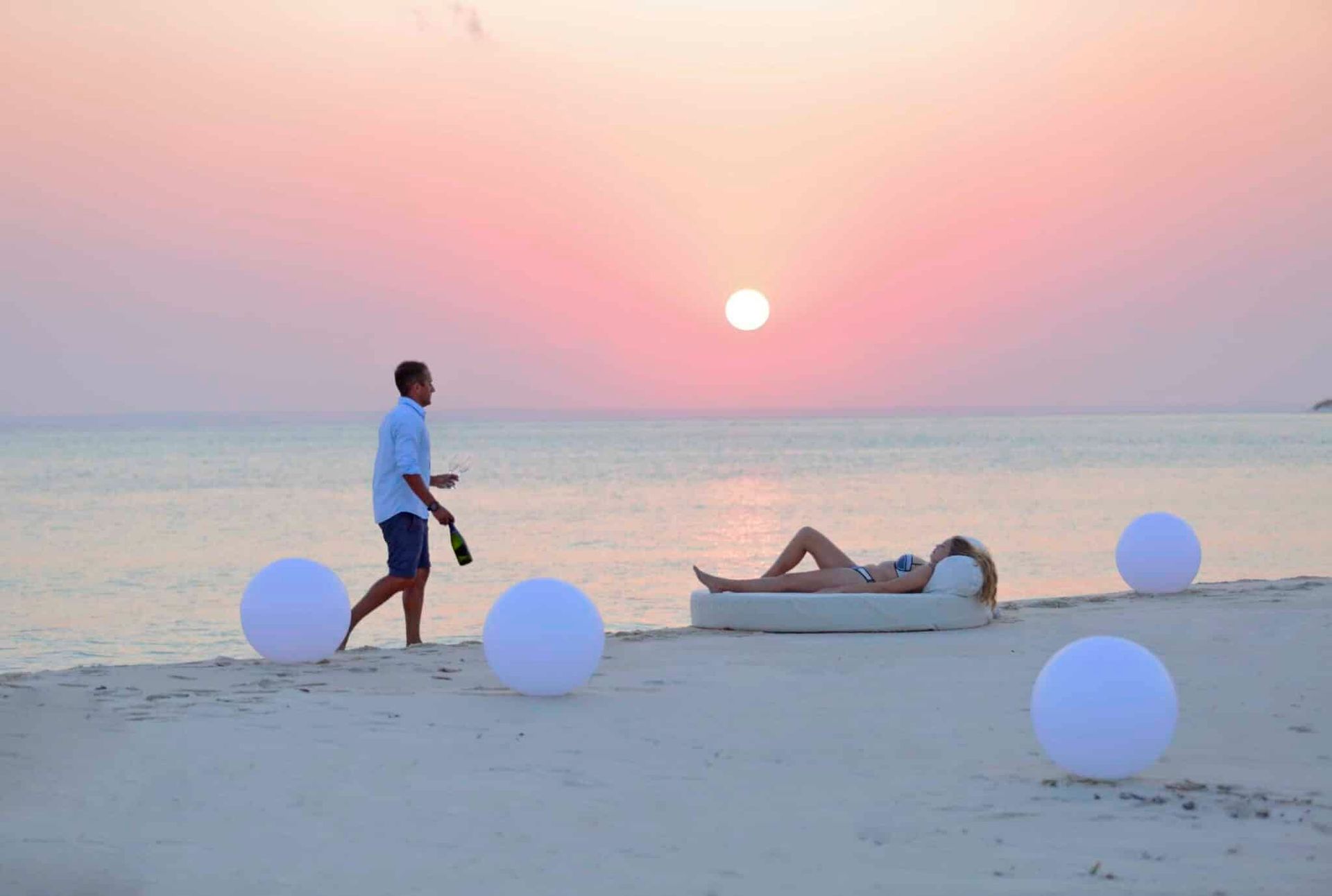 Two people on a beach at sunset, with white balloons or lights on the sand and calm ocean water behind them.