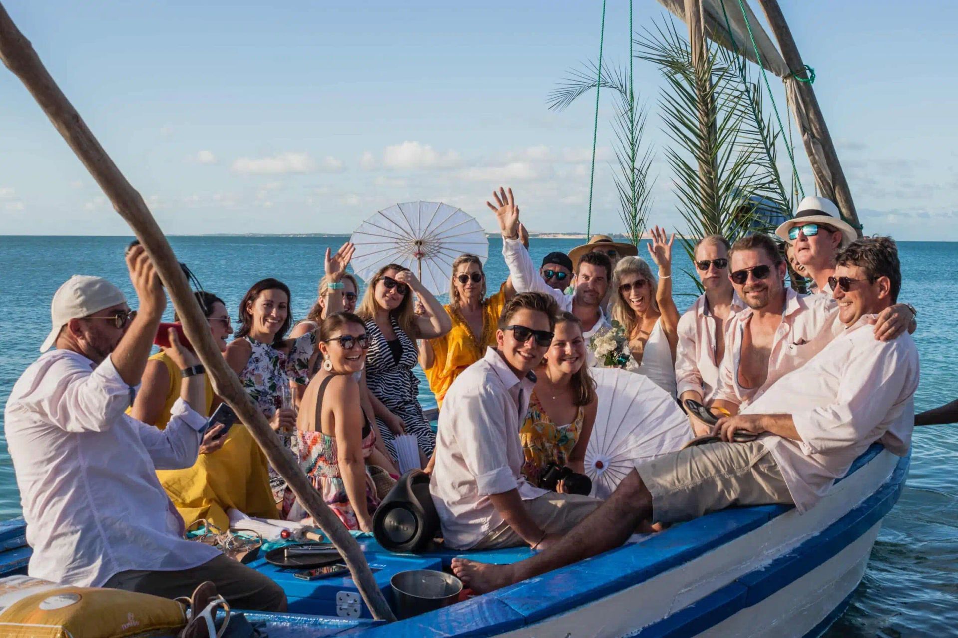 Group of people smiling on a blue-and-white boat at sea, waving with sailboat masts in the background