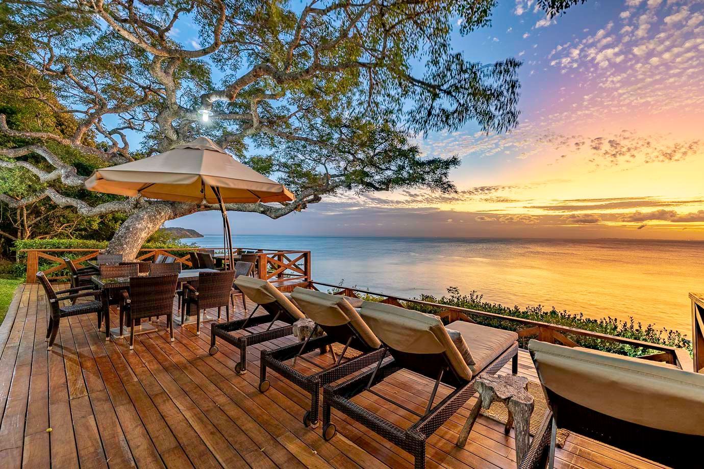 Sunset deck overlooking the ocean with lounge chairs, umbrella, and dining table under a tree