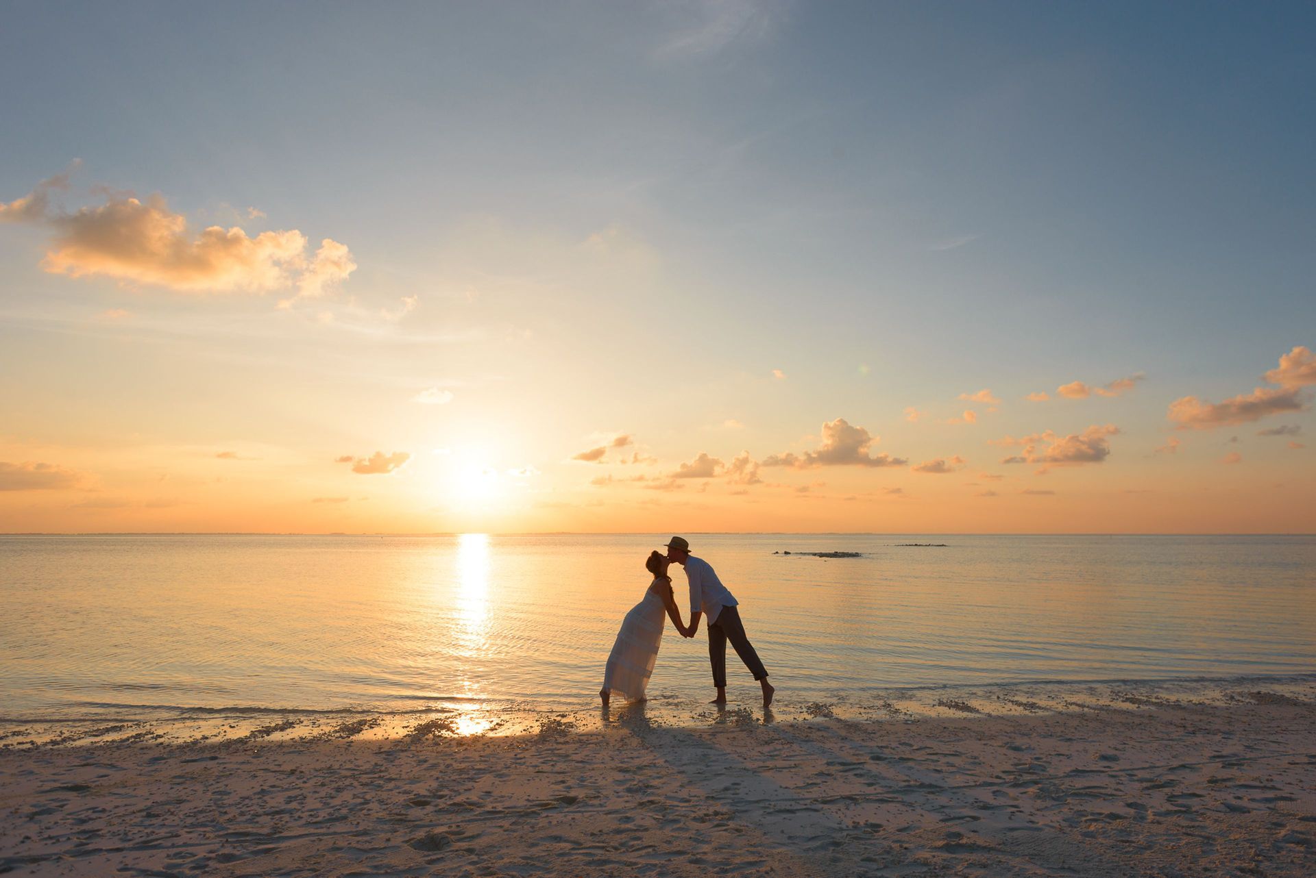 Two people dancing on a beach at sunset, with golden light reflecting on calm water.