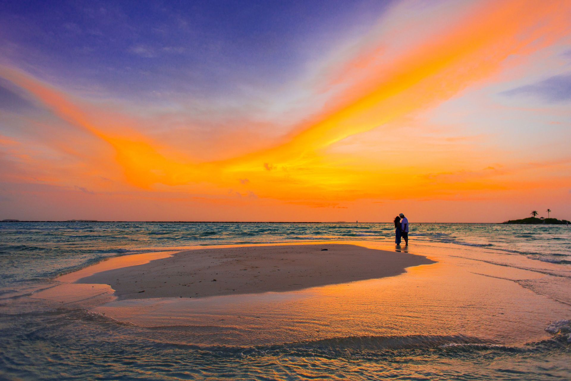 Two people standing on a sandy beach at sunset under a vivid orange and purple sky