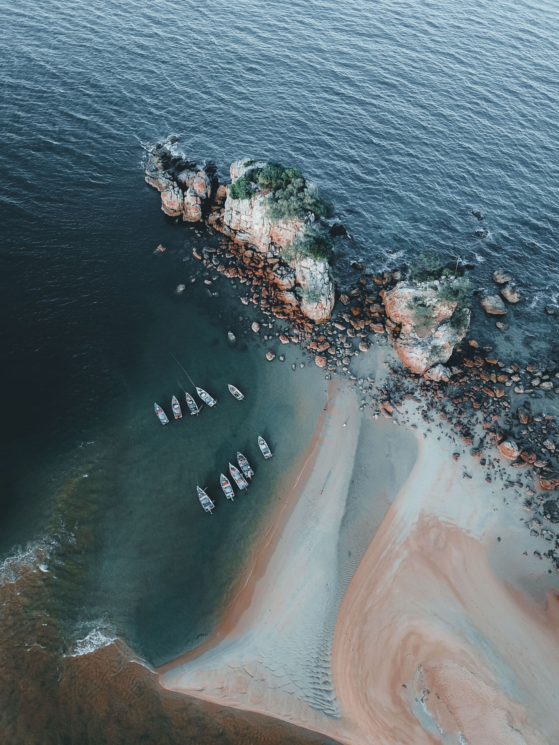 Aerial view of sailboats near a rocky shoreline with turquoise water and white sandbars