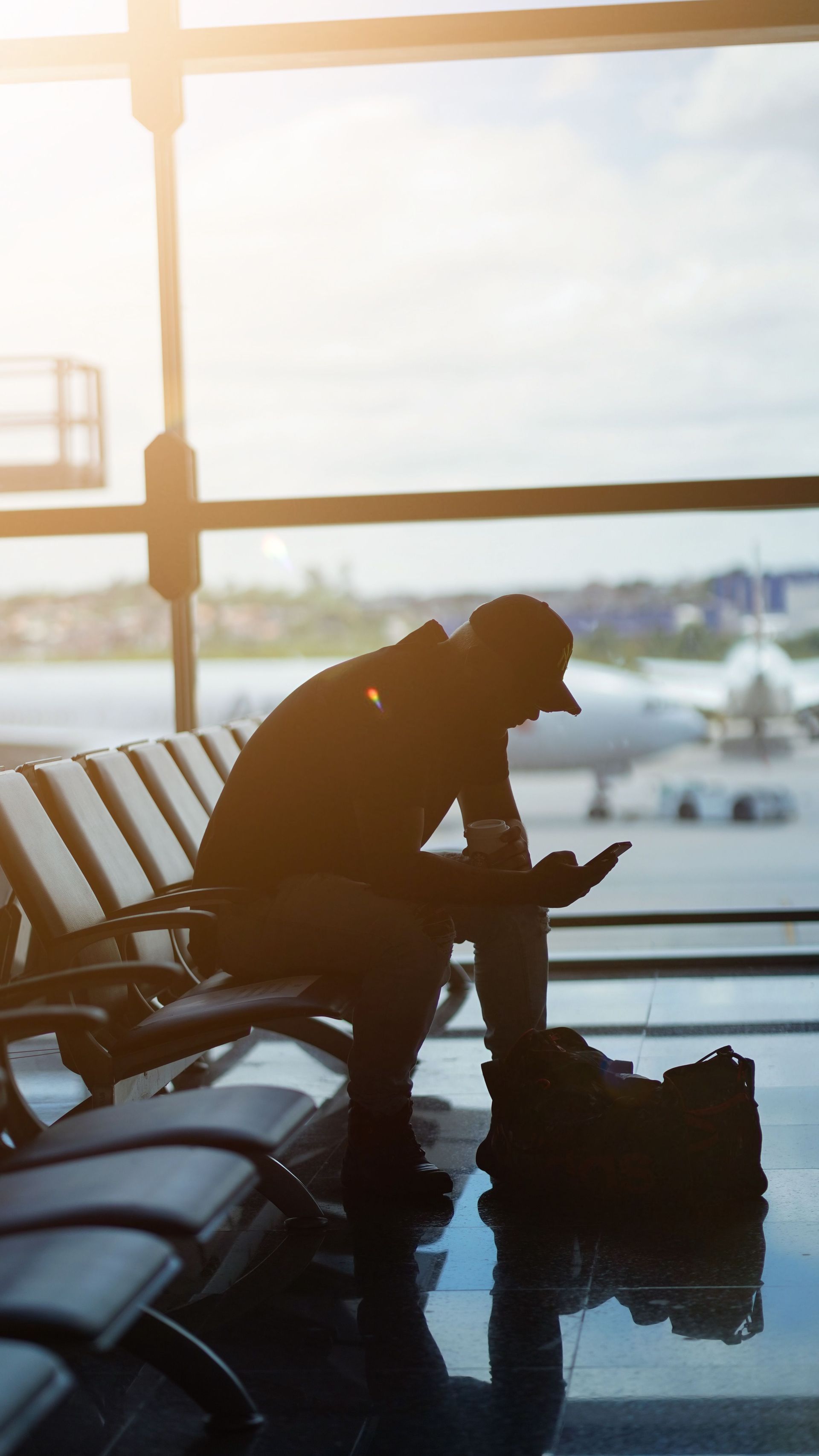 Silhouette of a traveler seated at an airport gate, looking at a phone beside luggage, with bright runway light behind.