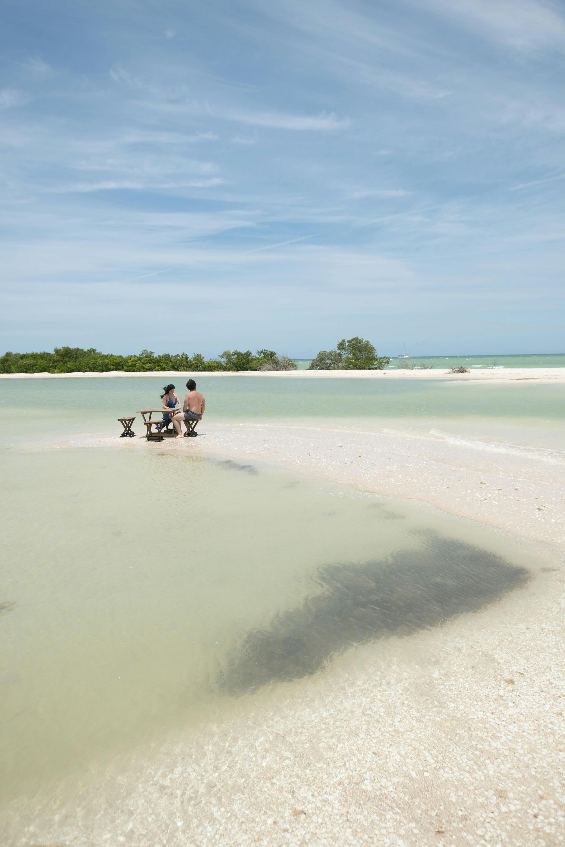 People sitting on a sandy tropical beach beside clear shallow water and a blue sky
