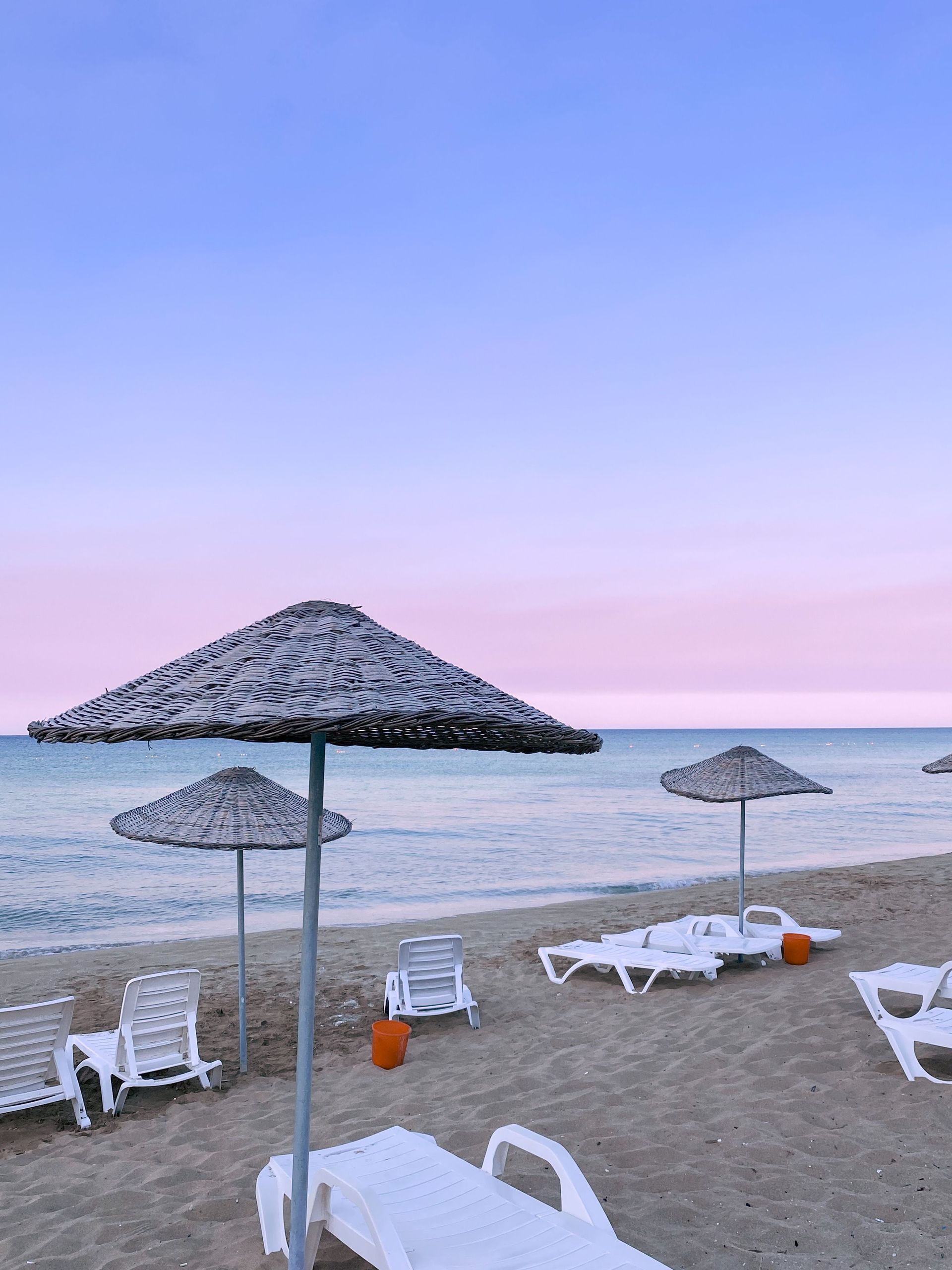 Beach chairs and straw umbrellas on a sandy shore at sunset, with calm sea and pastel sky.