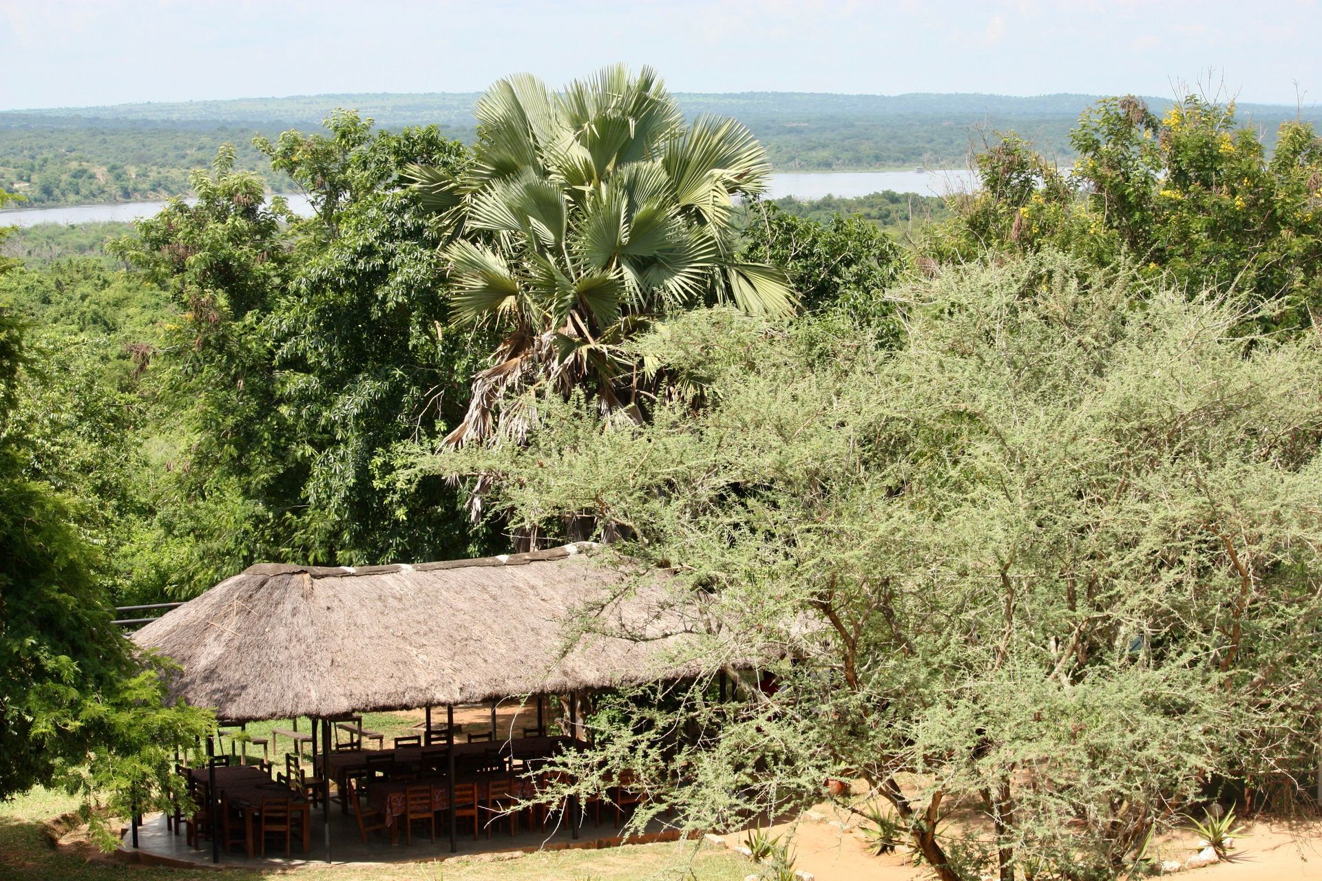Thatched hut amid lush green trees, overlooking a river or lake in the background.