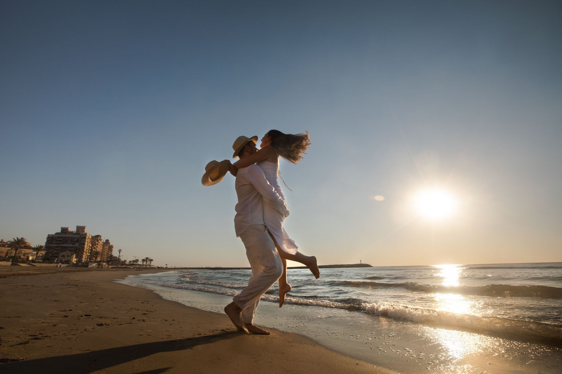 Couple dancing on a sunlit beach at sunset, with waves and a low sun in the background