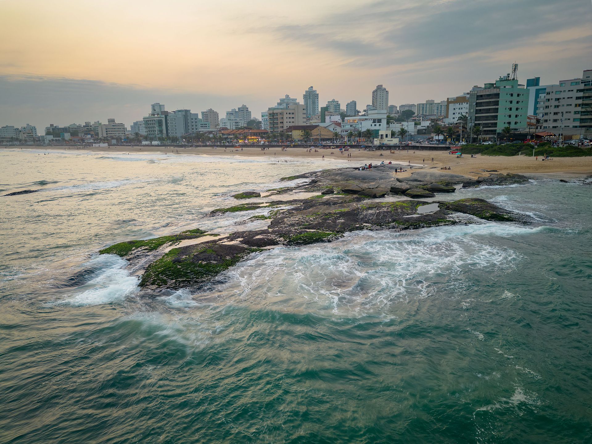 Rocky shoreline with waves, a sandy beach, and a distant coastal city skyline at sunset