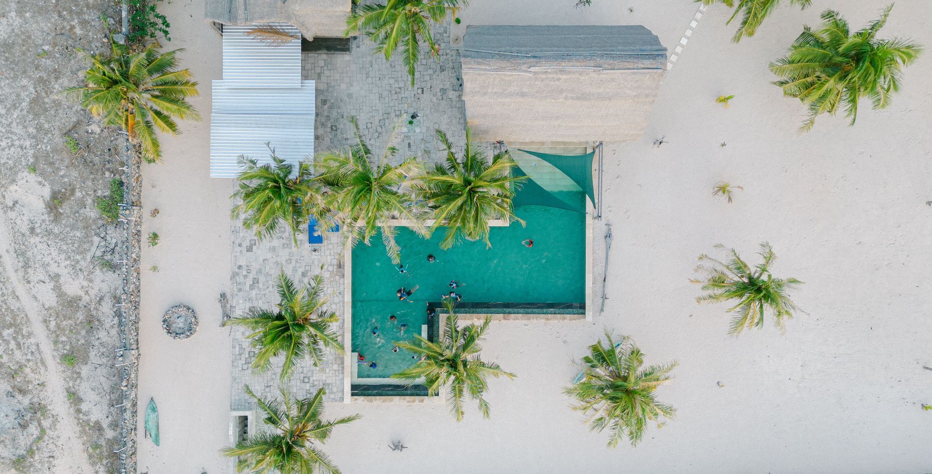 Aerial view of a turquoise swimming pool surrounded by palm trees and sandy ground.