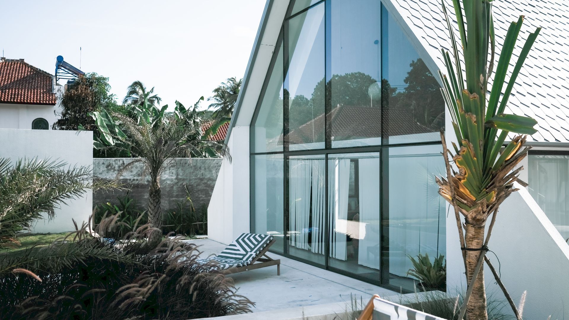 Modern glass-walled house with patio seating and tropical landscaping