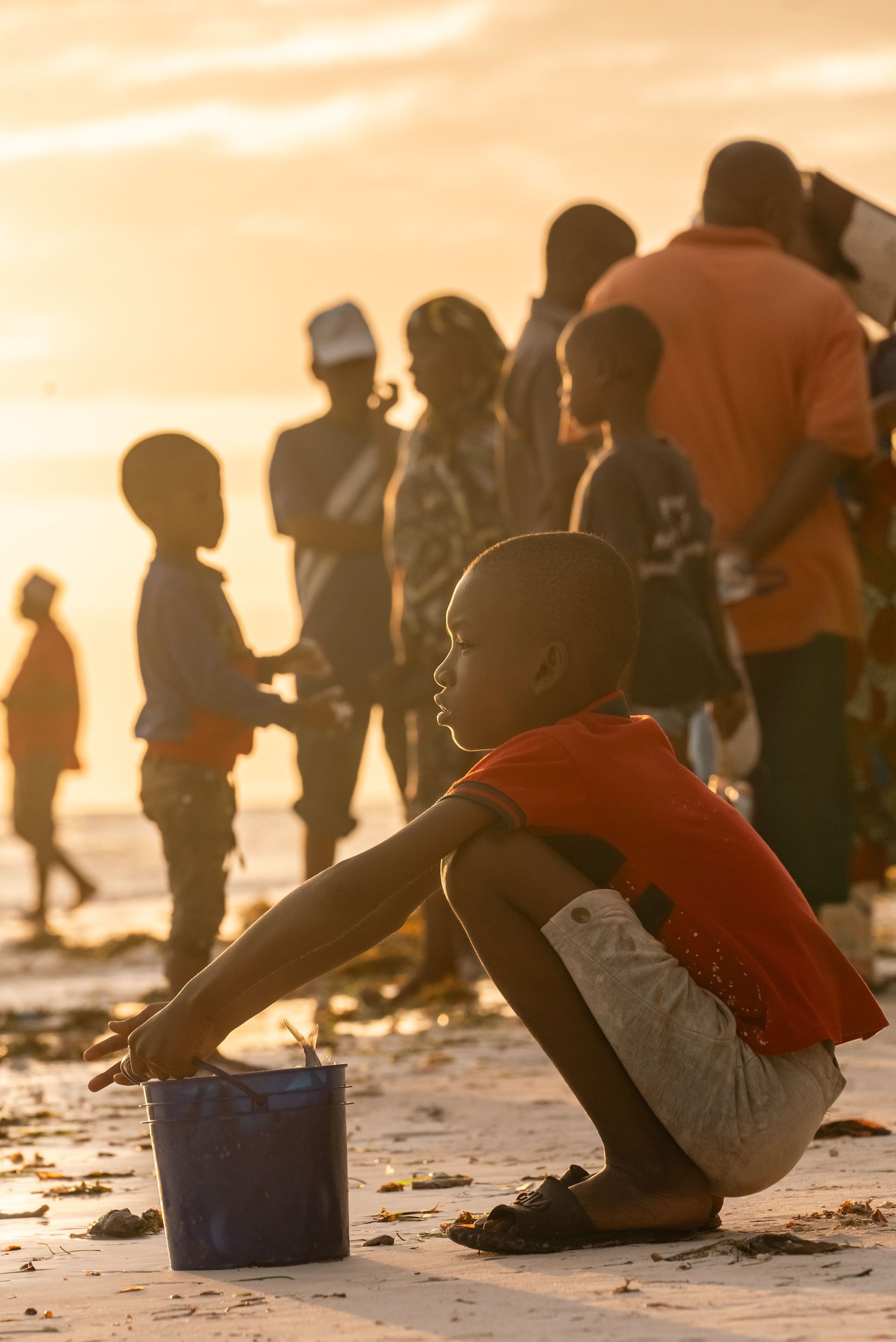 Children playing on a beach at sunset, one crouching by a blue bucket in the foreground.