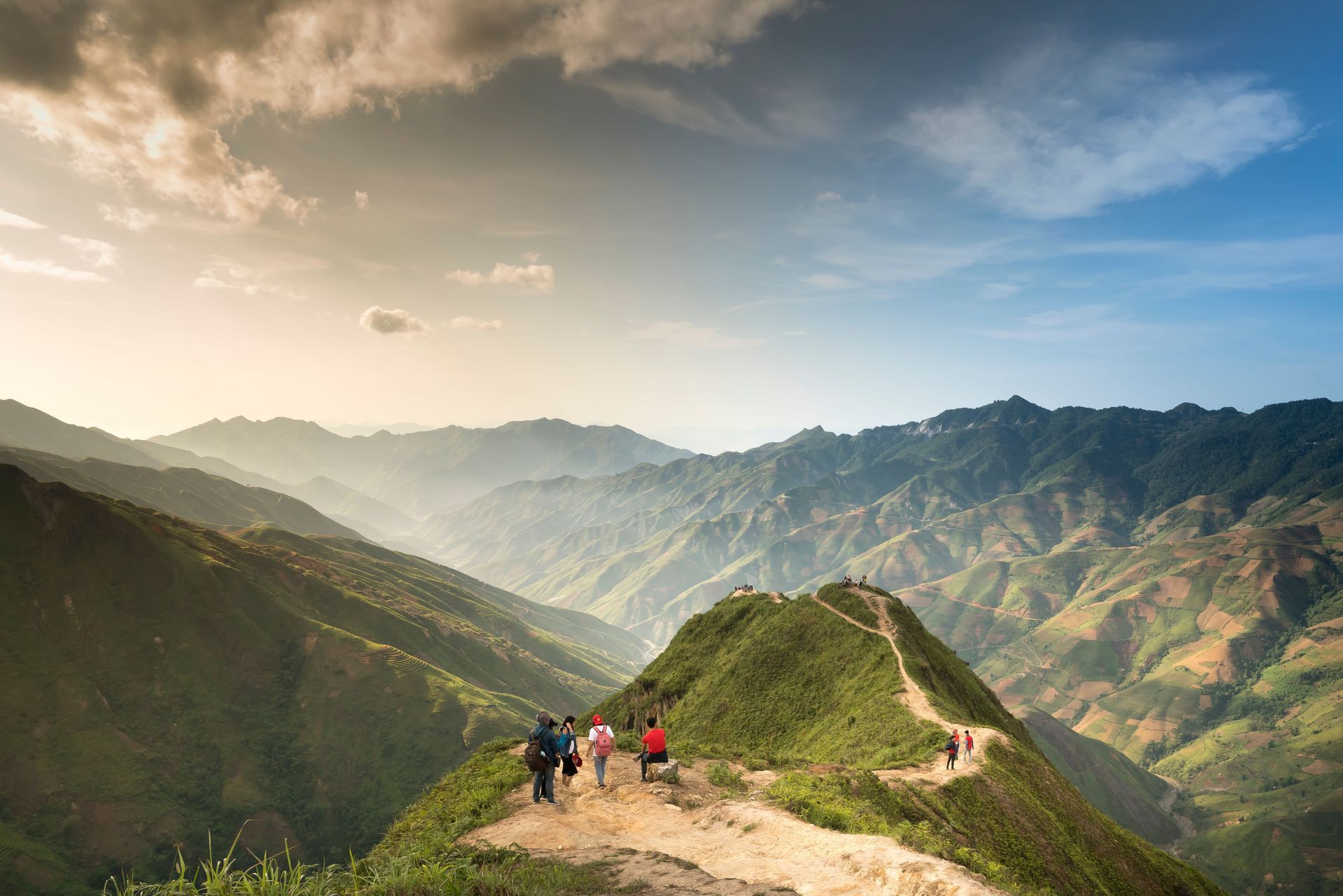 Hikers on a narrow mountain trail overlooking a green valley and sunlit peaks