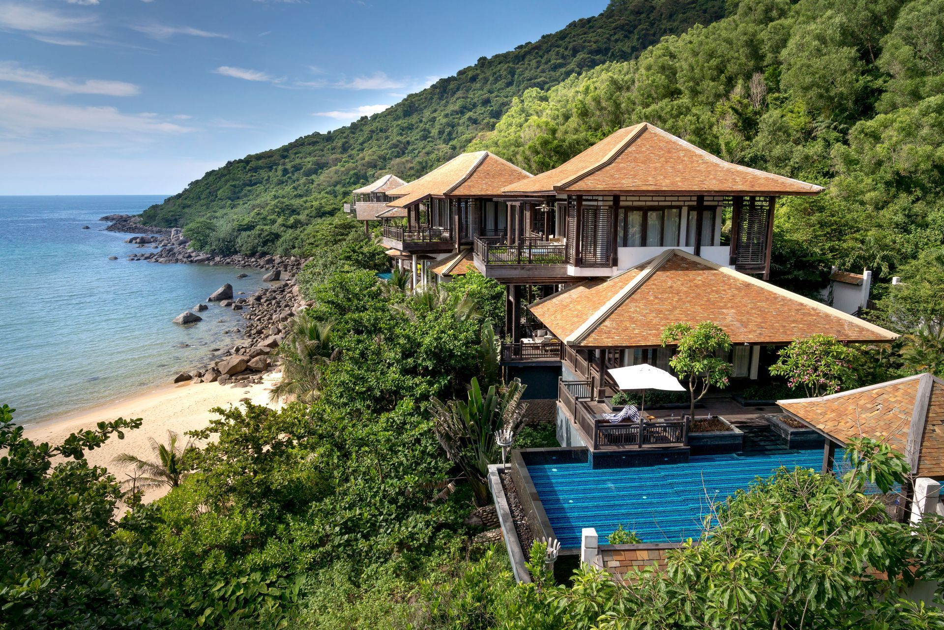Seaside villa with orange roofs, pool, and lush greenery overlooking a rocky beach and blue sea