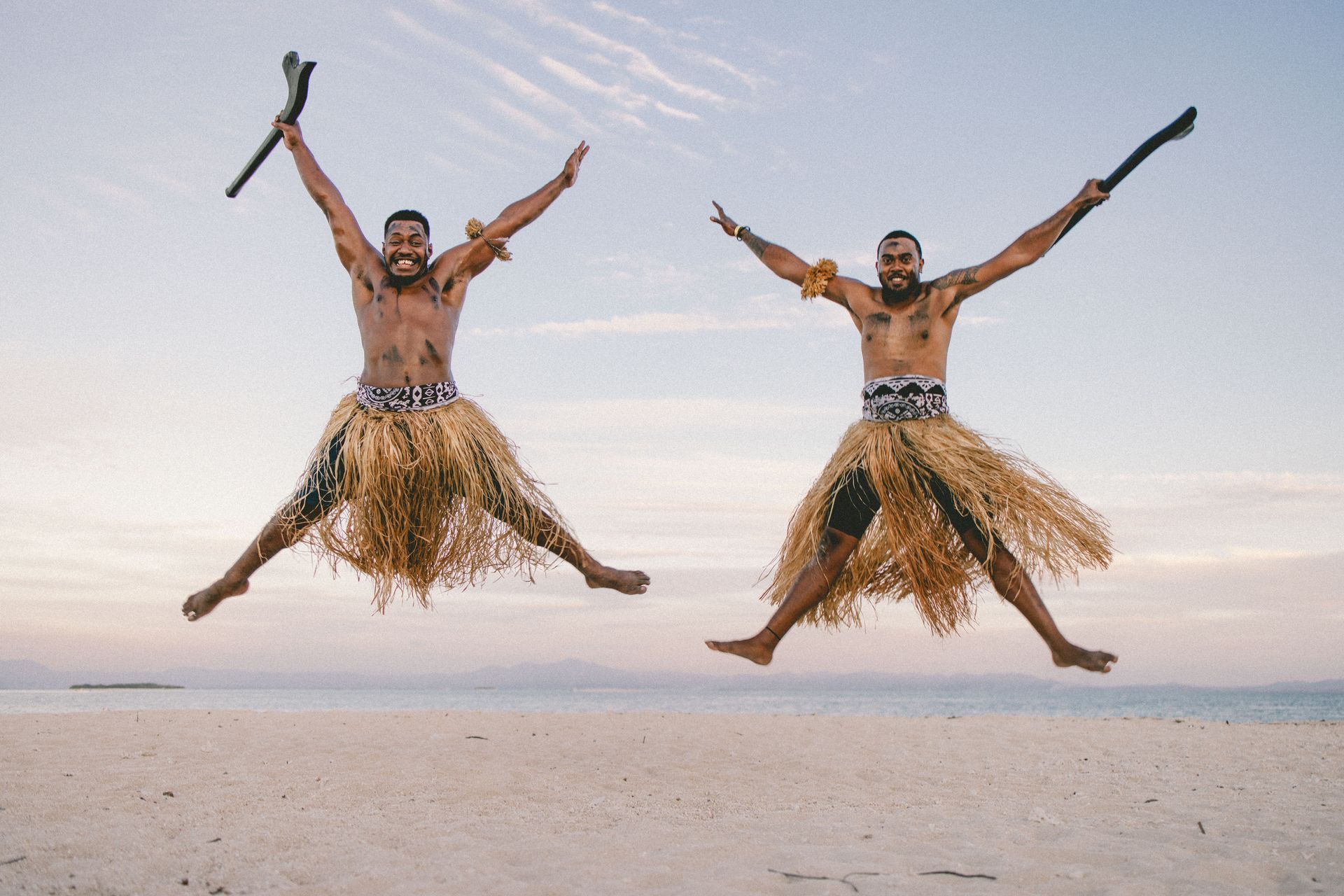 Two men in grass skirts leap on a beach at sunset, holding clubs overhead.