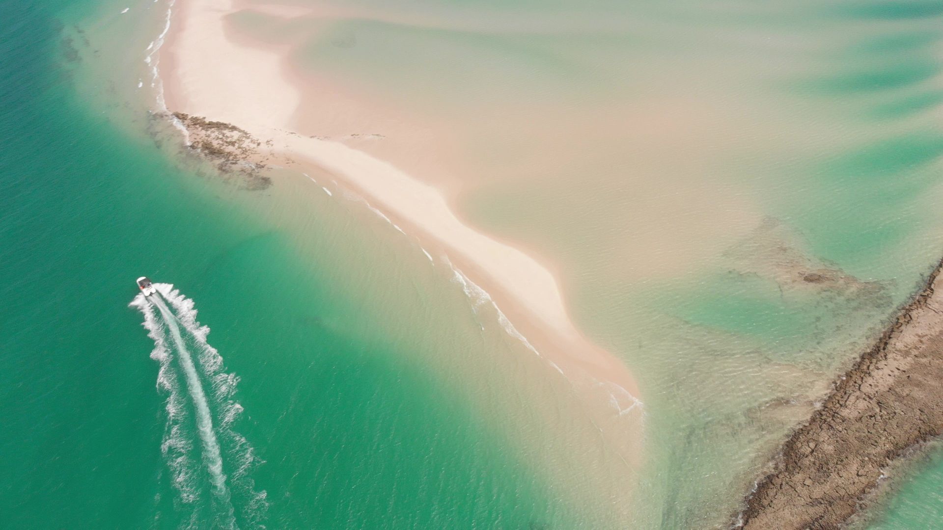 Aerial view of a turquoise bay with a speedboat leaving a white wake near a sandy beach and rocky shoreline