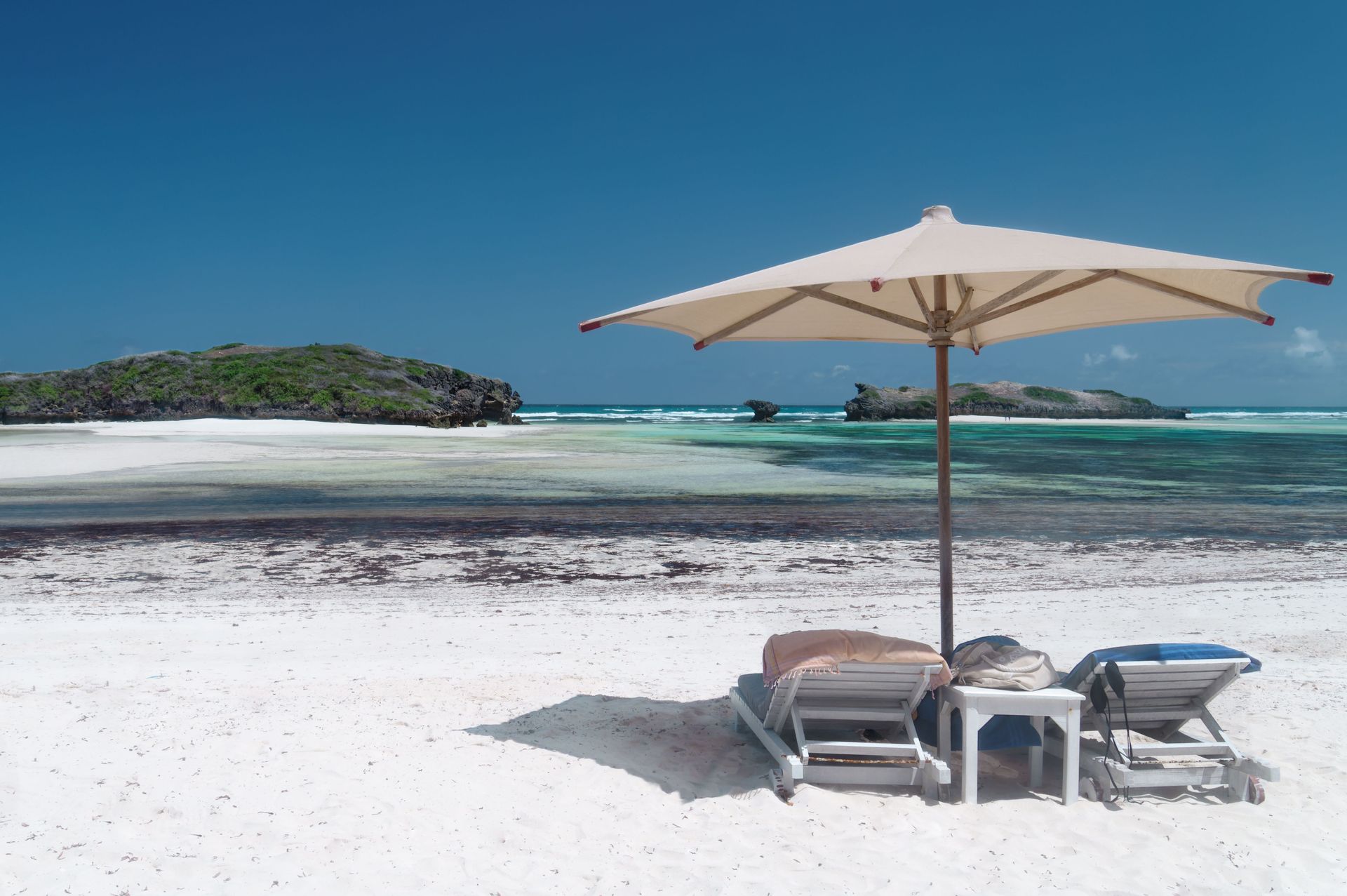 Beach chairs under a white umbrella on a sunny tropical shore with turquoise water and an island in the distance