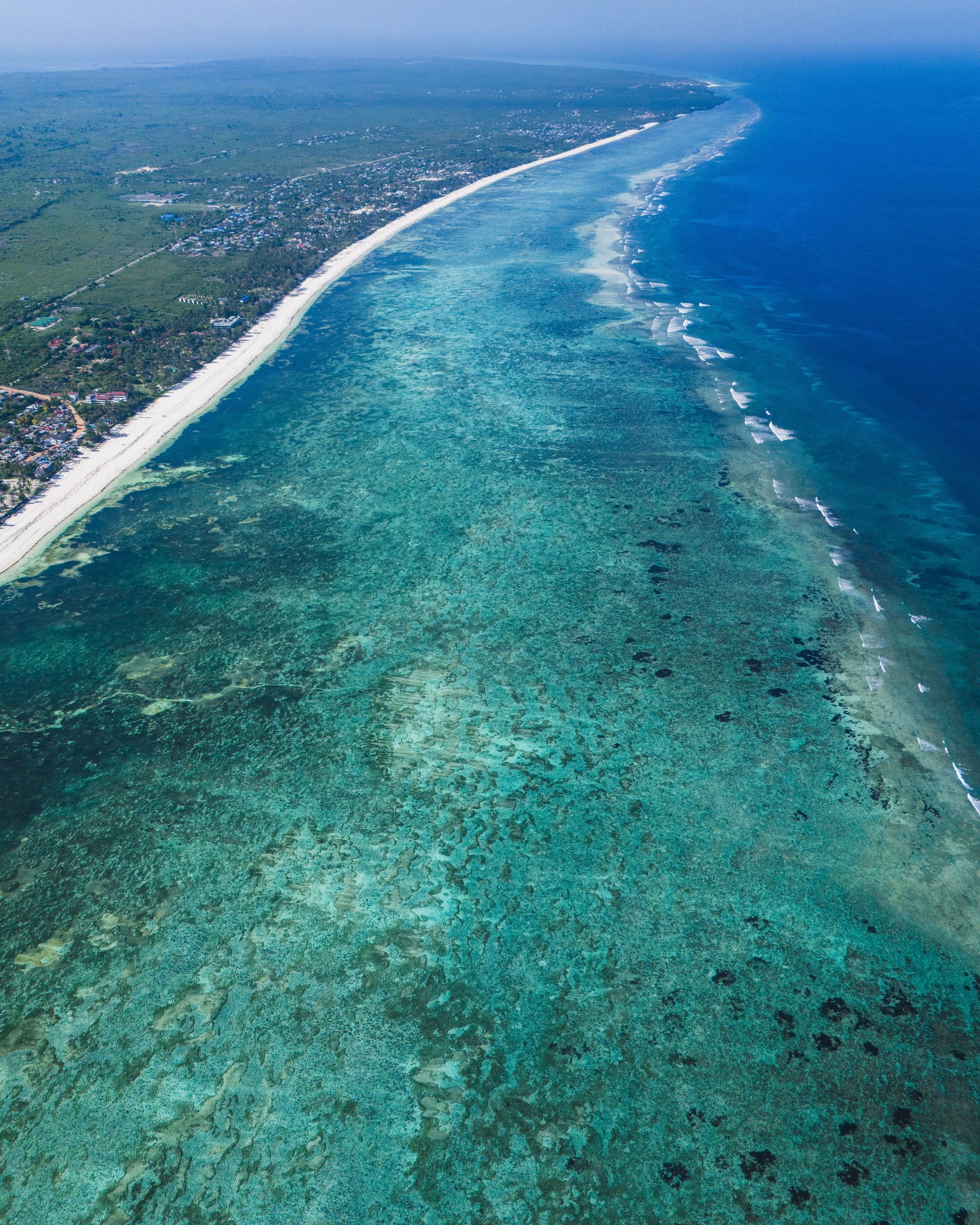 Aerial view of a turquoise coral reef beside a sandy coastline and blue ocean