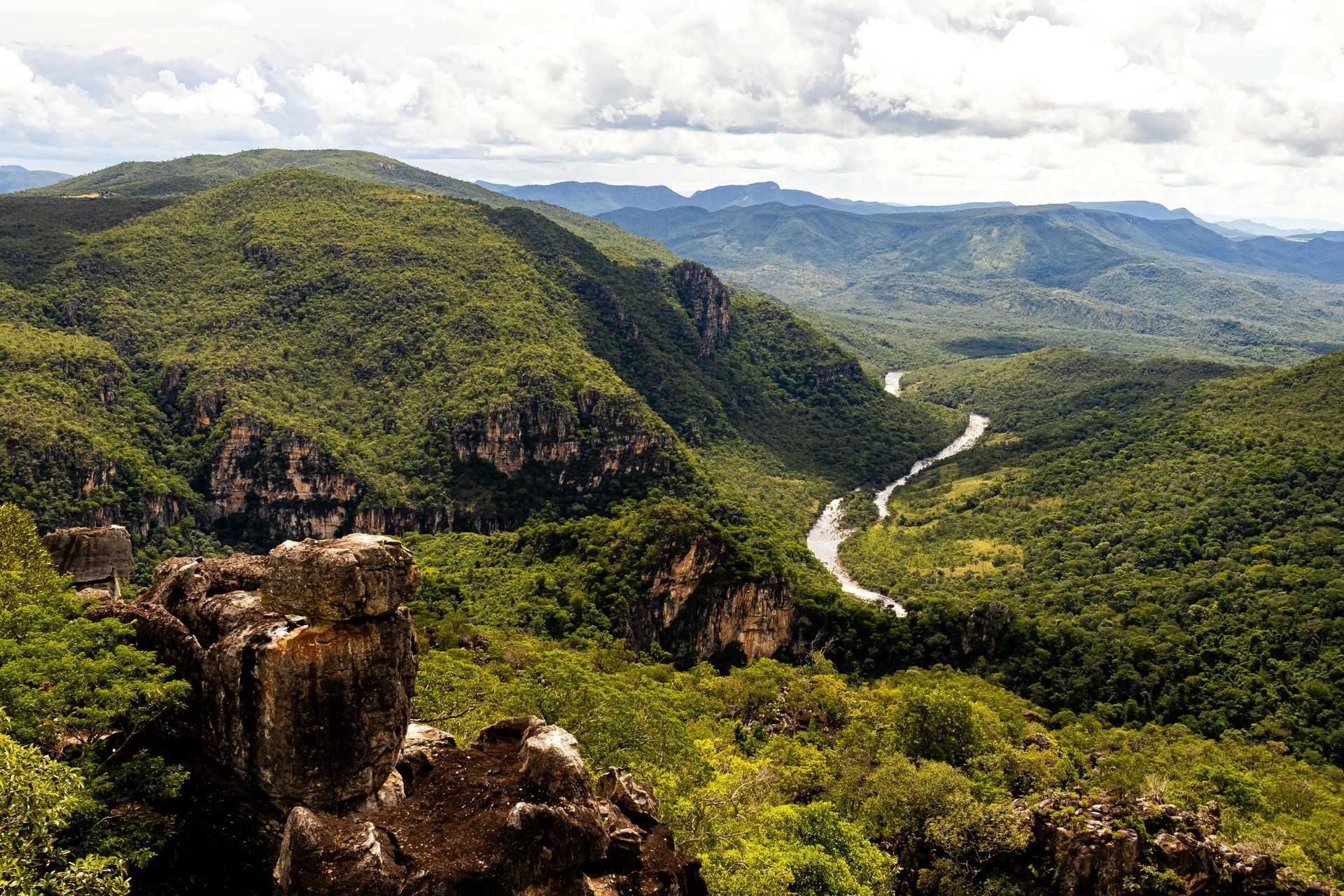 Green mountainous valley with a winding river and rocky foreground under a cloudy sky