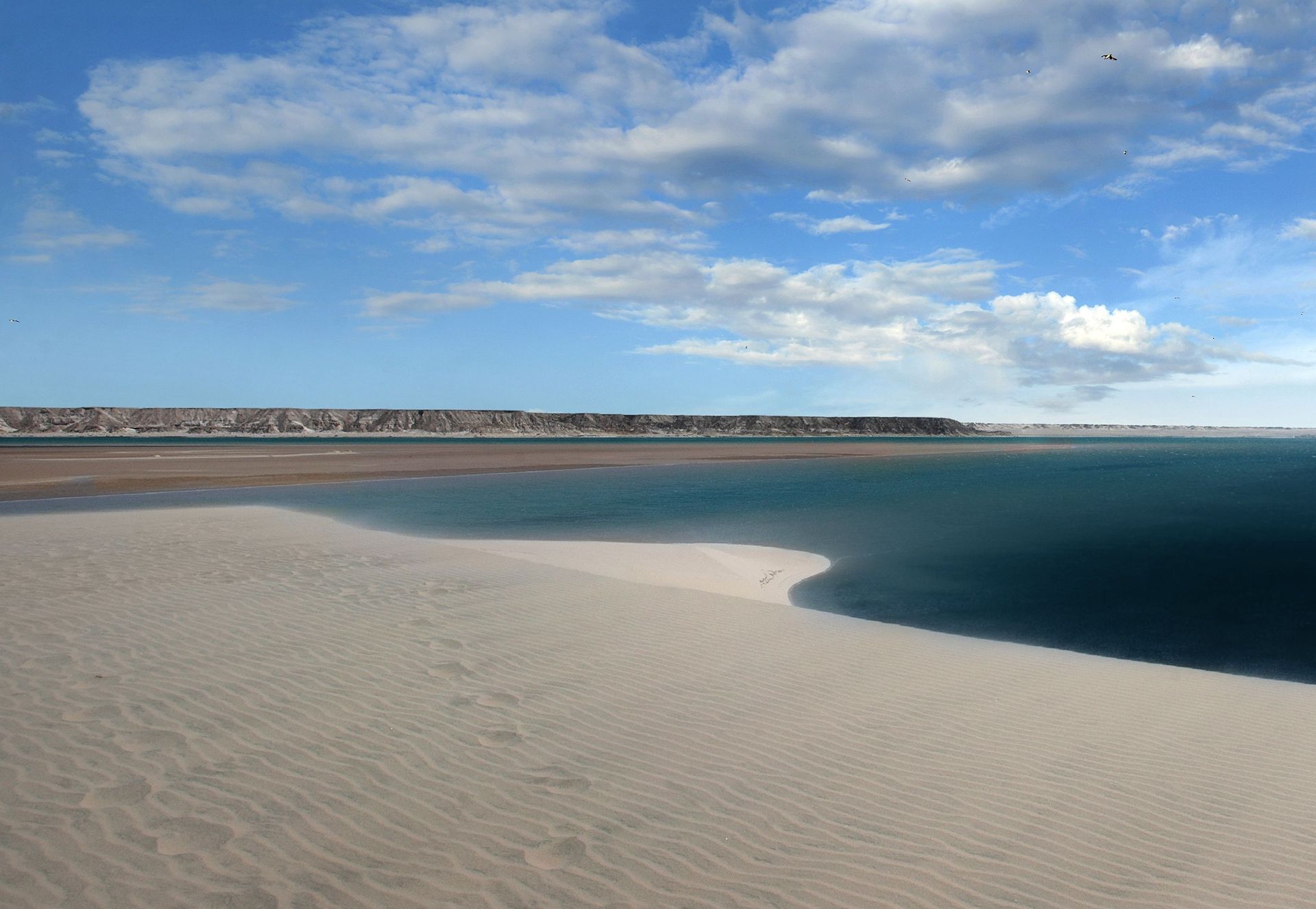 Sandy beach curving beside calm blue water under a partly cloudy sky