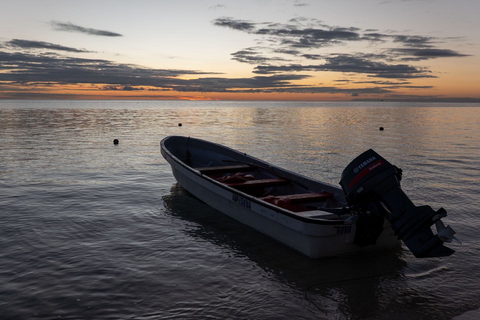 Small motorboat floating on calm water at sunset, silhouetted against a cloudy sky.