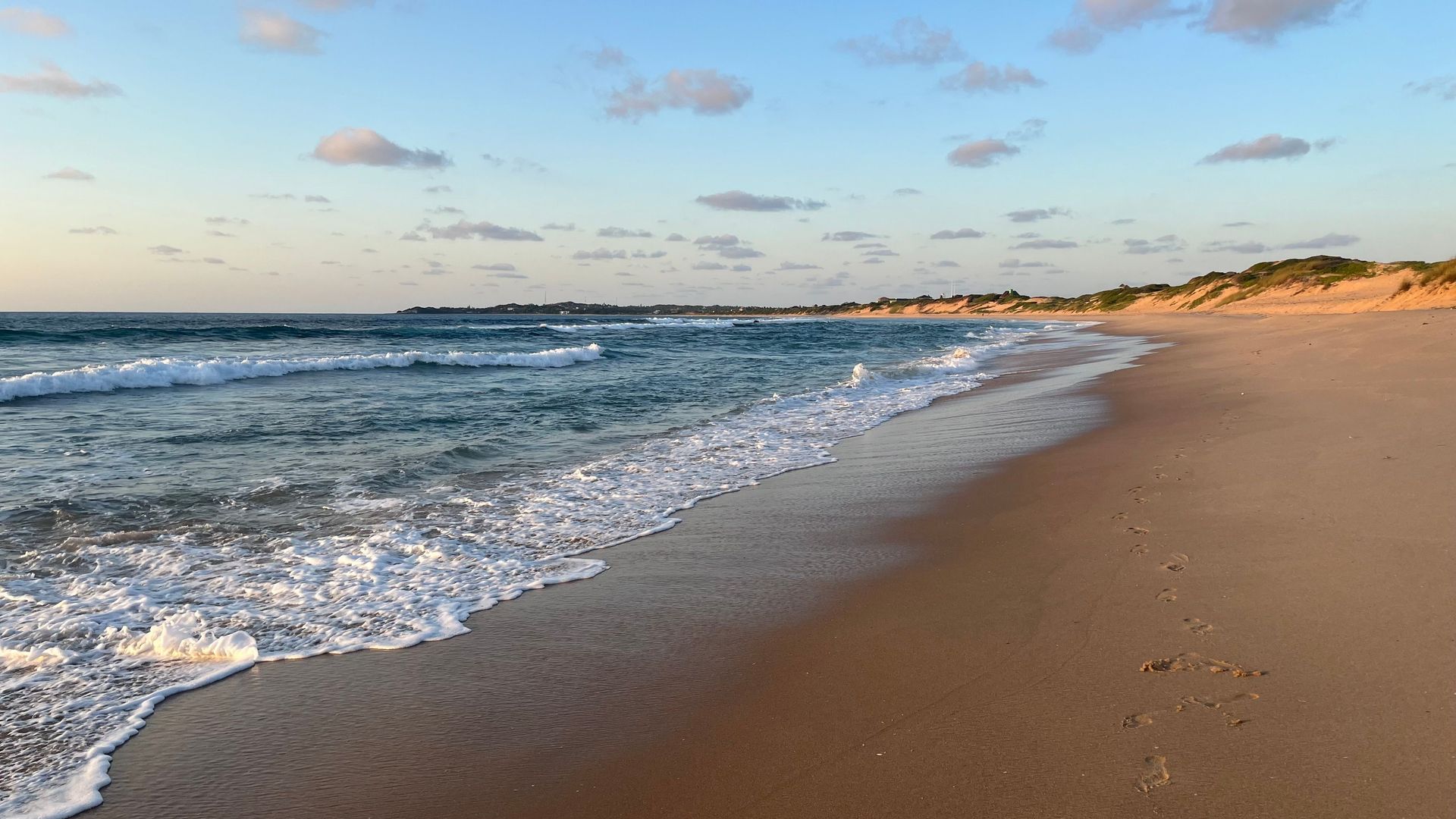 Sunny beach shoreline with gentle waves, sandy shore, and a partly cloudy sky at sunset