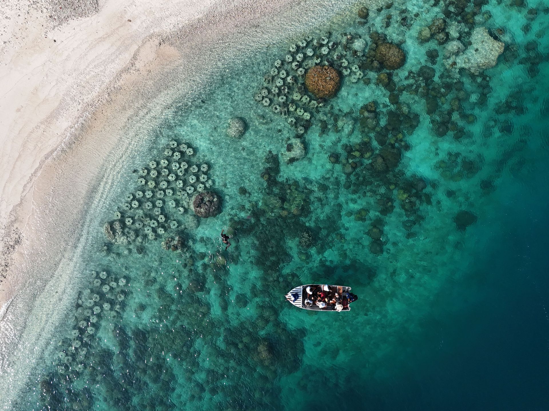 Aerial view of a small boat on turquoise water beside a white sandy shoreline and coral reef formations