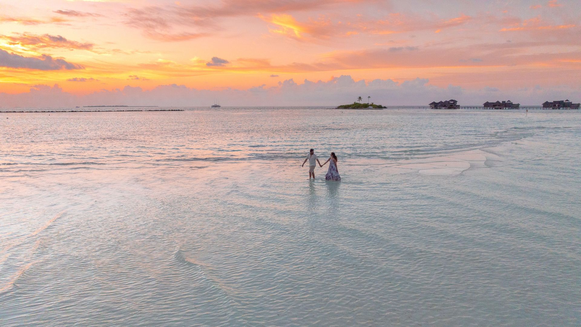 Two people wading in calm ocean water at sunset, with pink-orange clouds over the horizon.