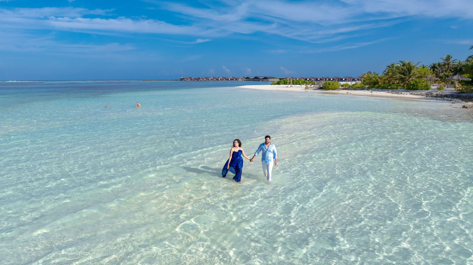 Two people standing in clear turquoise water near a sandy tropical beach under a bright blue sky