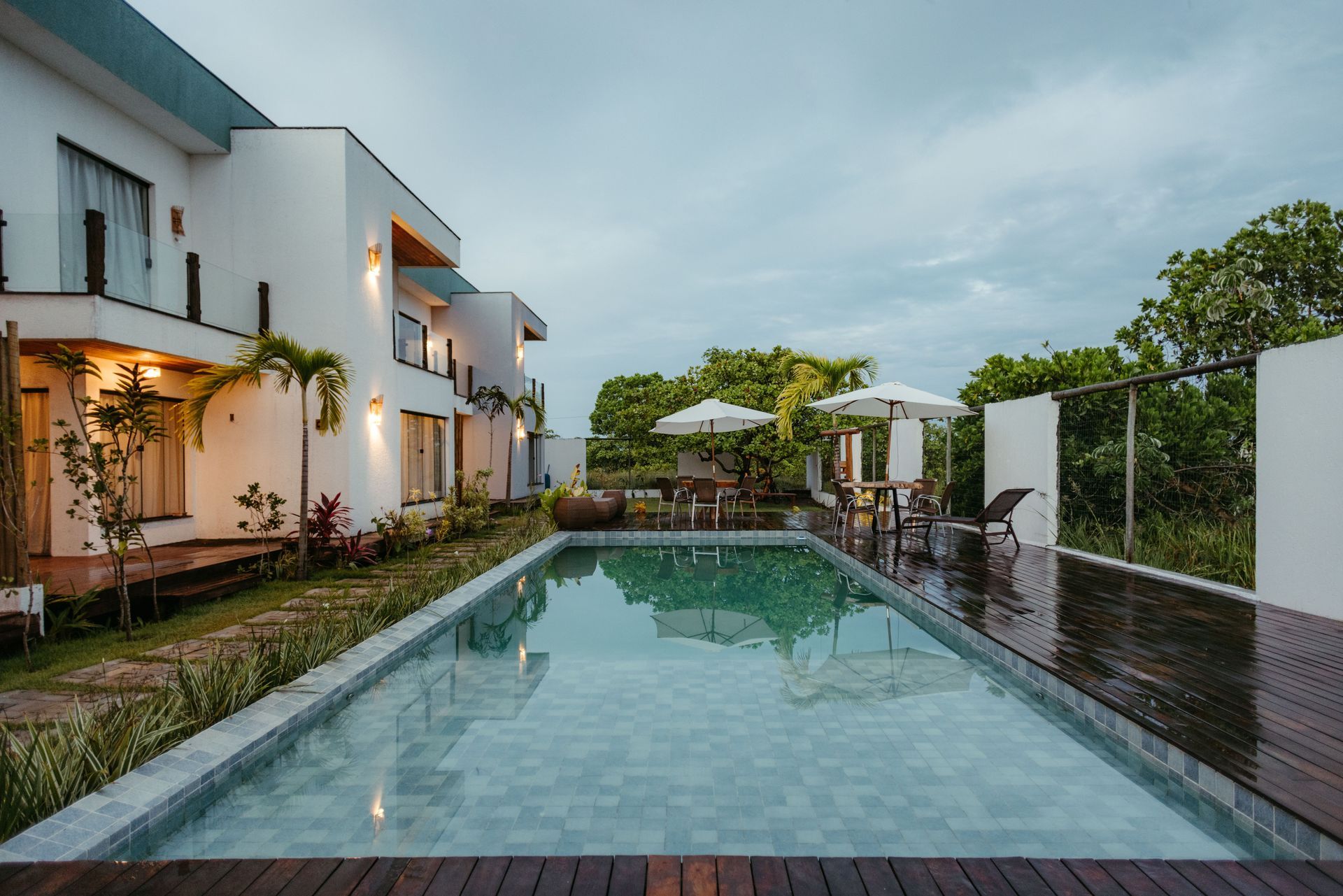 Modern white villa beside a long pool with wooden deck and lush greenery under cloudy sky