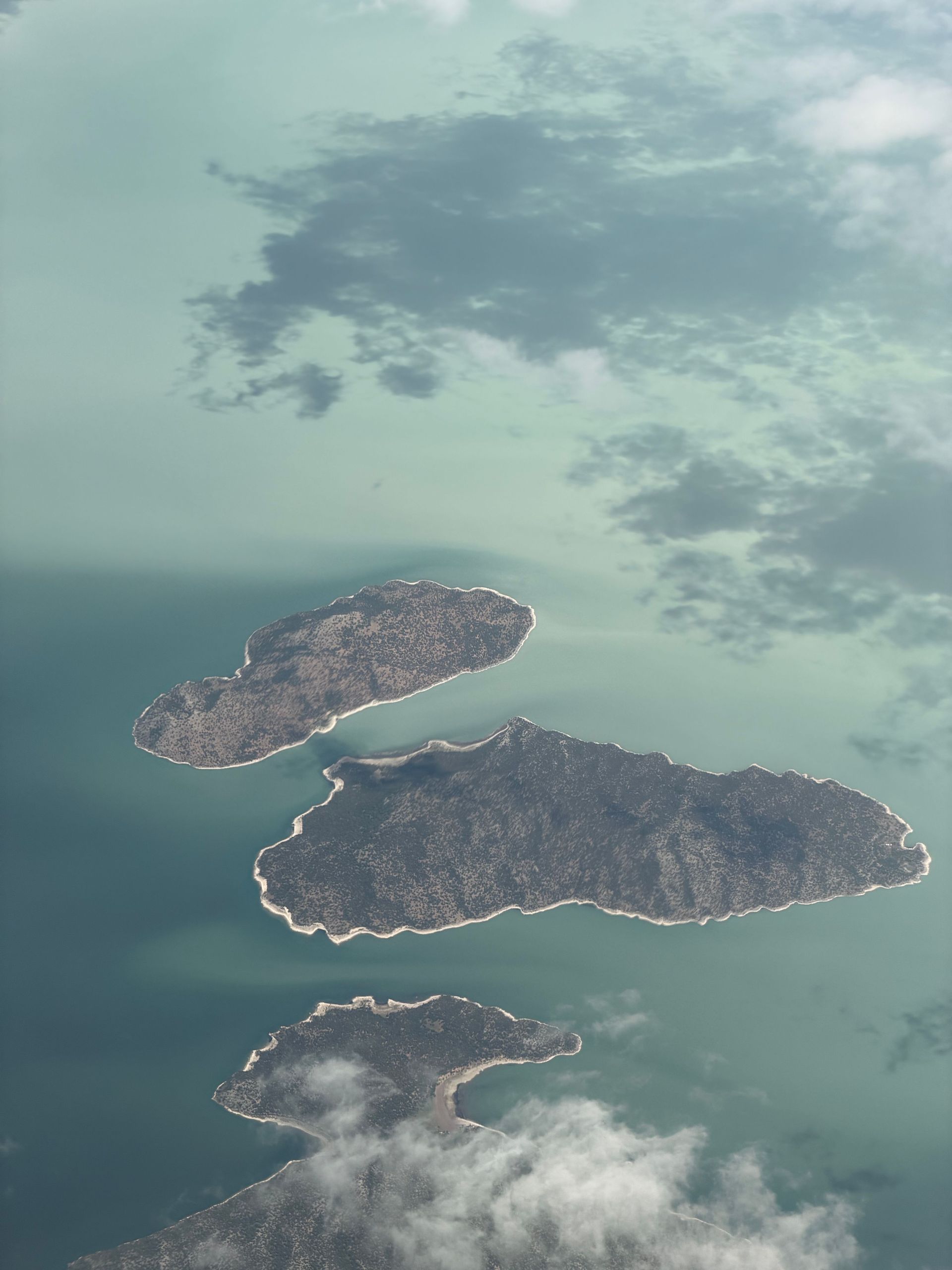 Aerial view of three small green islands surrounded by calm blue-green water and clouds.