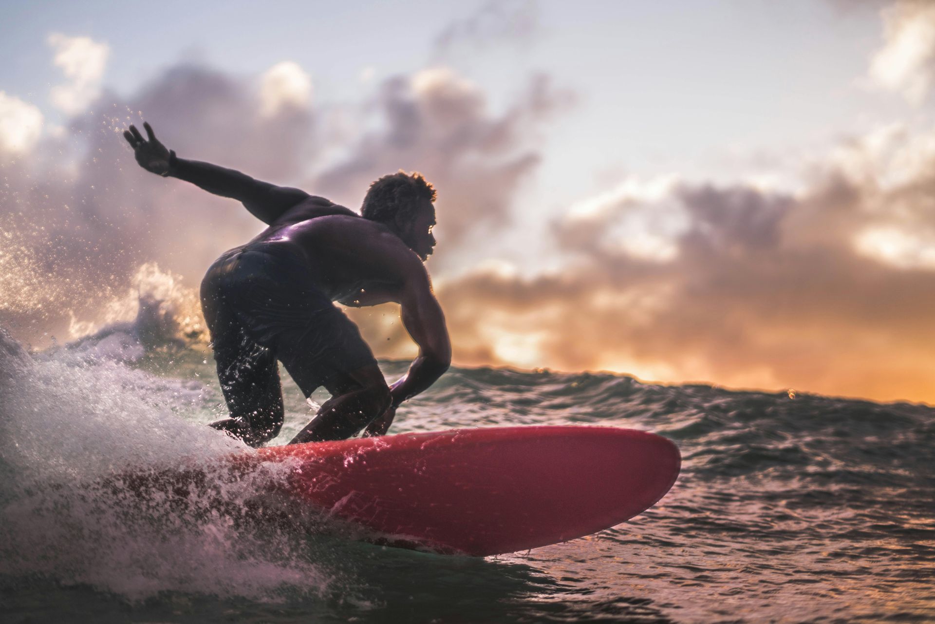 Surfer riding a red board on a wave at sunset, with water spray and a cloudy sky.