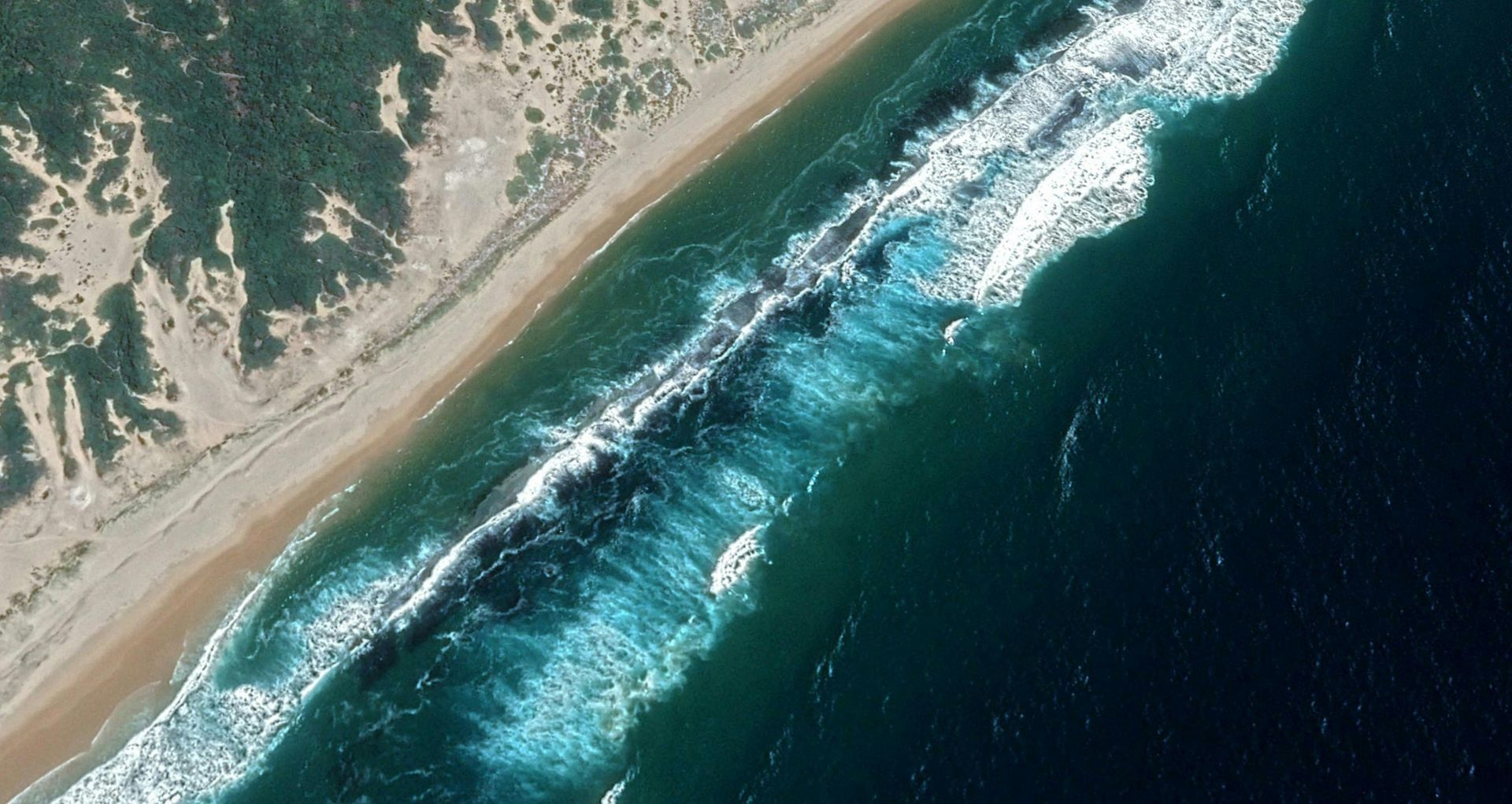 Aerial view of a sandy beach with teal waves breaking along a rocky coastline.