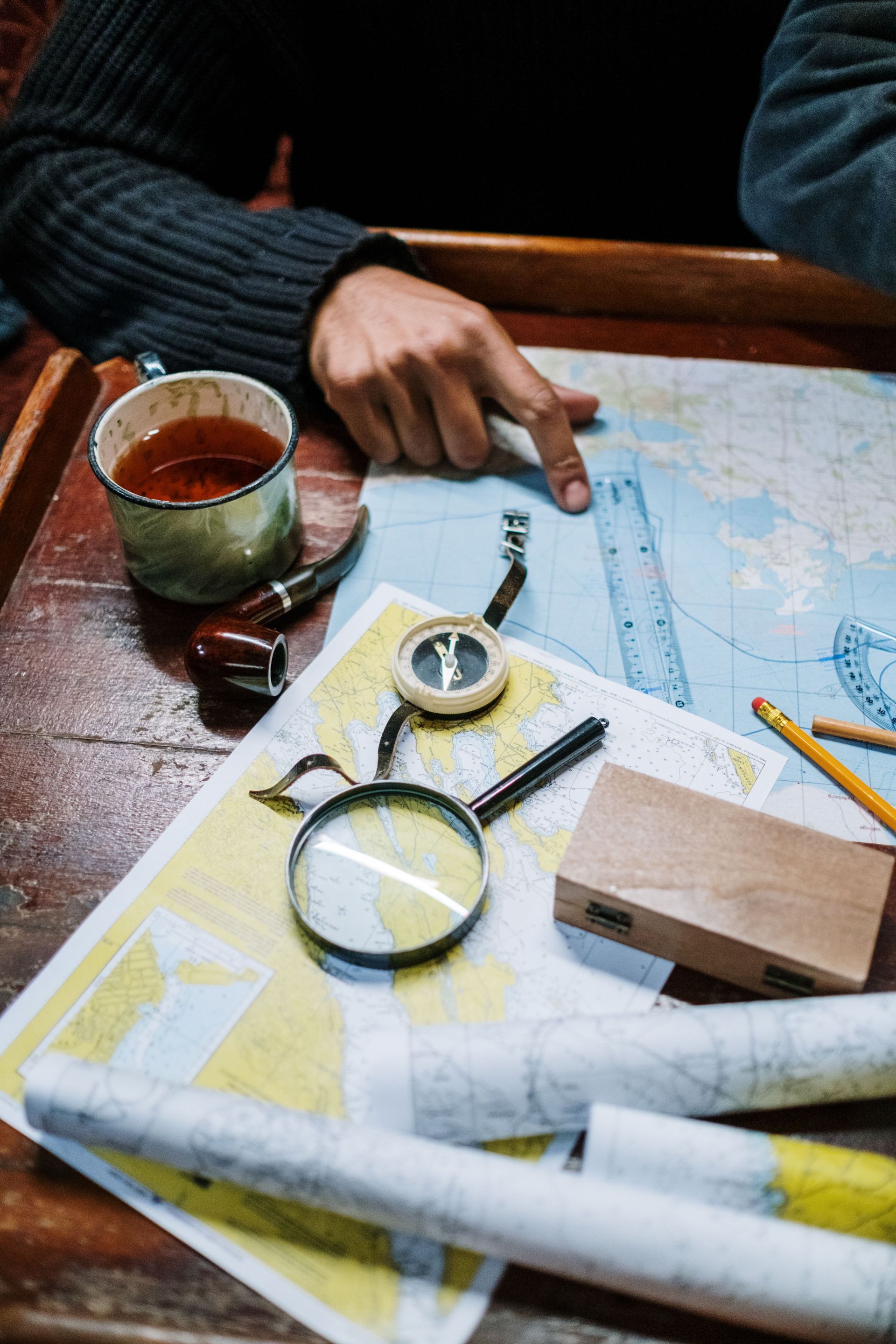 Hands pointing at a map on a table with a compass, magnifying glass, cup, and rolled maps