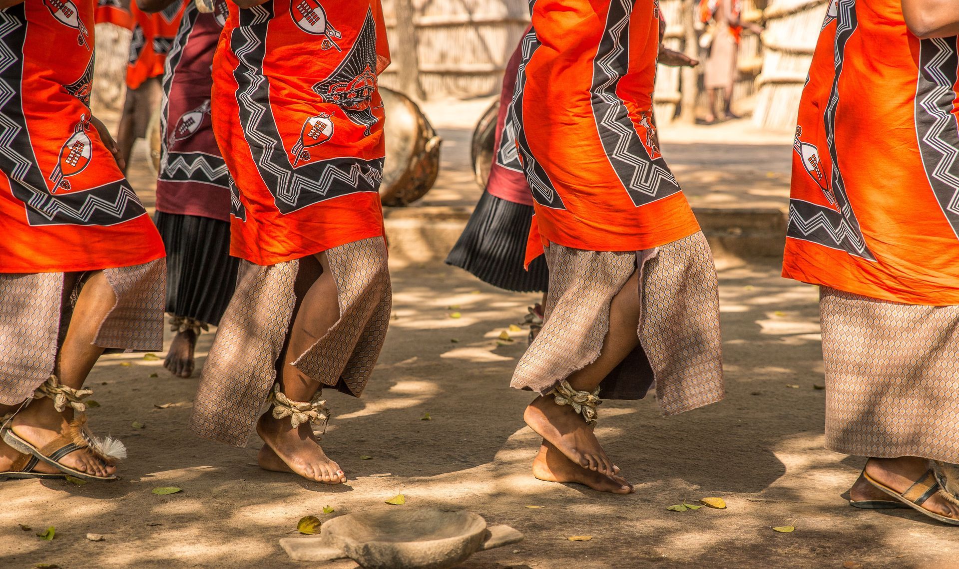 People in orange and red garments walking on a dusty street, seen from the waist down.